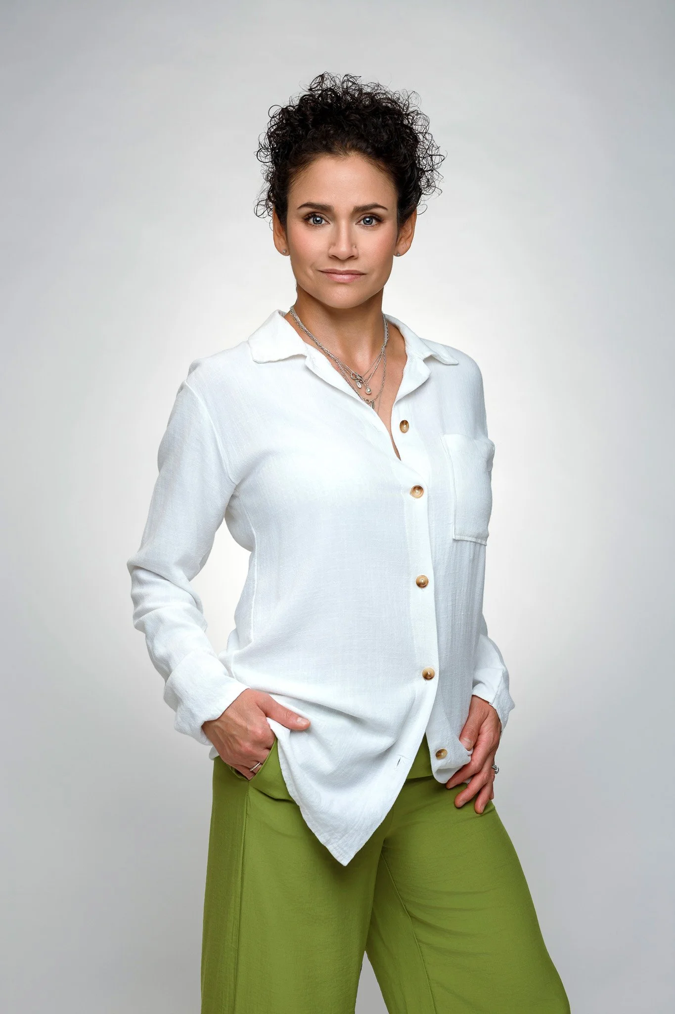 A woman with short curly dark hair wearing a white button-up shirt and green pants.