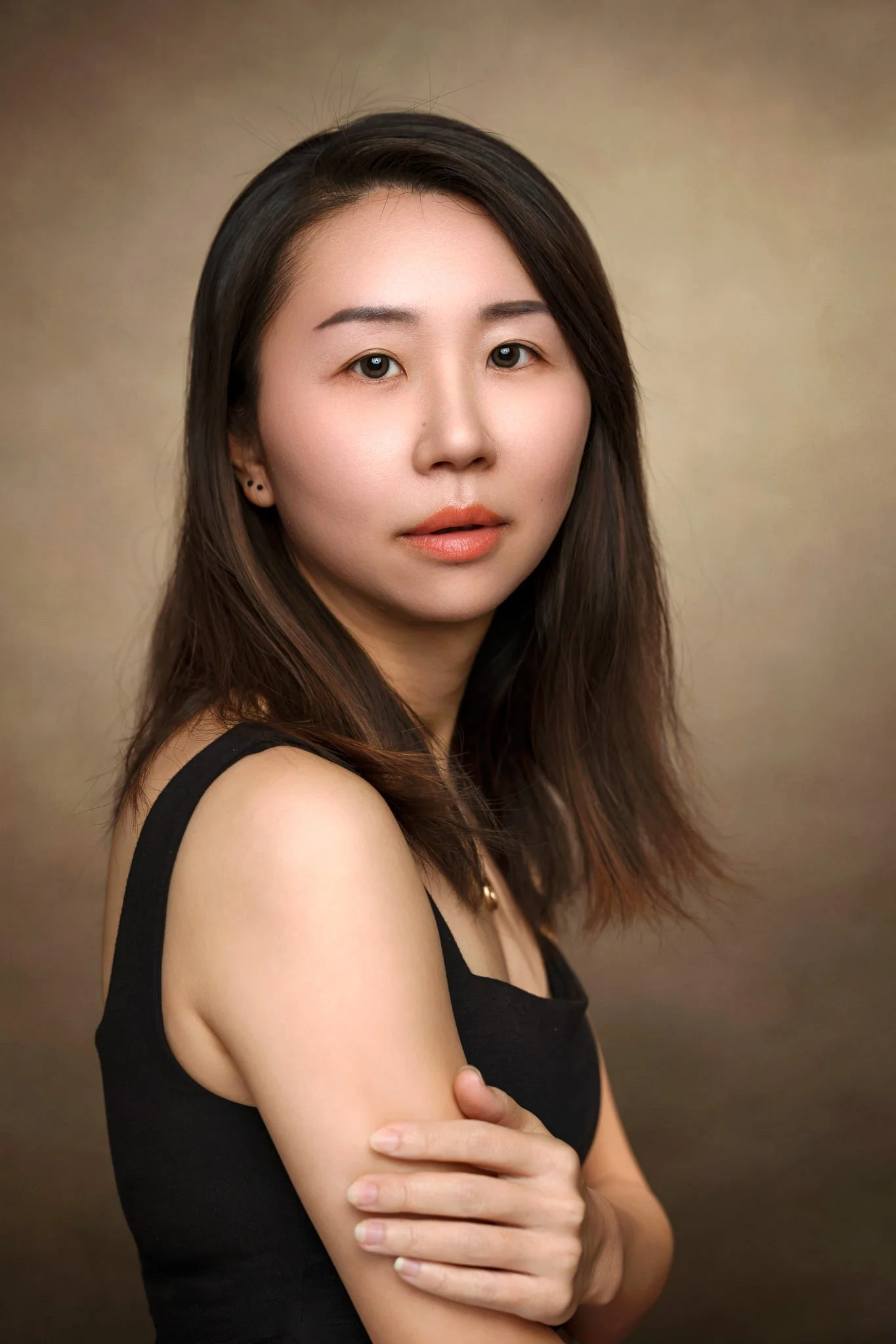 A woman with long dark brown hair and light skin, wearing a black sleeveless top, standing against a beige background with her right arm crossed over her stomach and her left arm resting on her right arm.