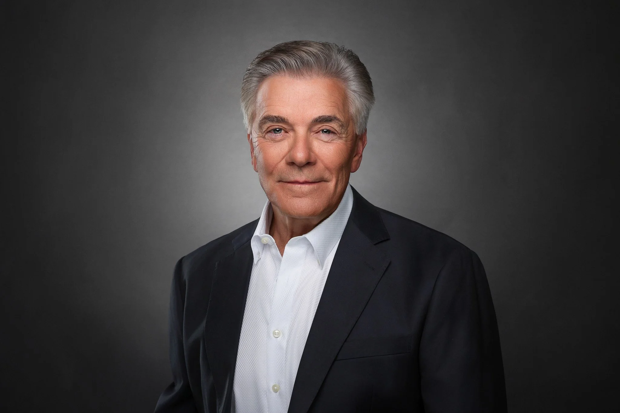 An older man with gray hair wearing a black suit and white dress shirt, smiling softly against a dark gray background.
