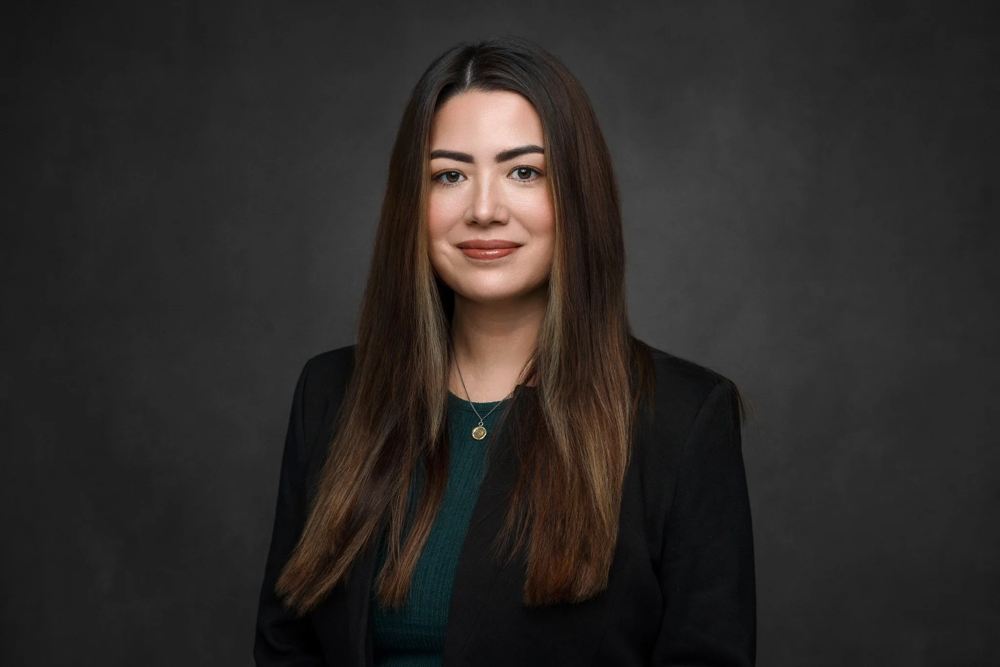 Portrait of a woman with long brown hair, wearing a black blazer and a gold necklace, against a dark gray background.