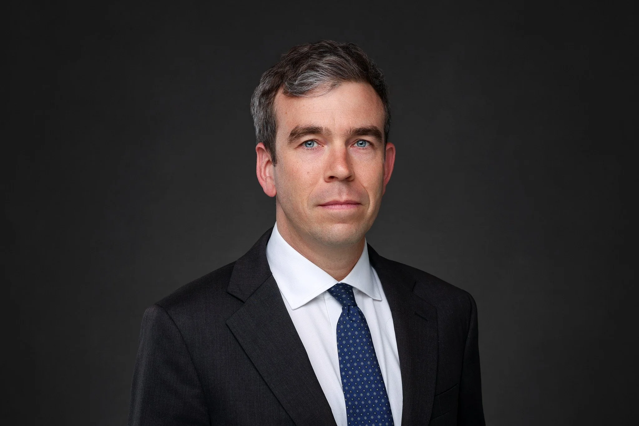 A professional headshot of a man with short dark hair and blue eyes, wearing a dark suit, white shirt, and blue tie with small patterns, against a dark gray background.