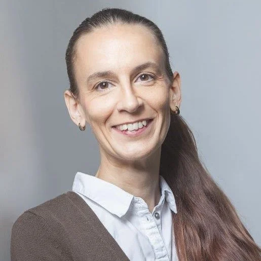 Porträt einer lächelnden Frau mit braunem Haar, grauem Hintergrund, weißem Hemd und brauner Jacke.