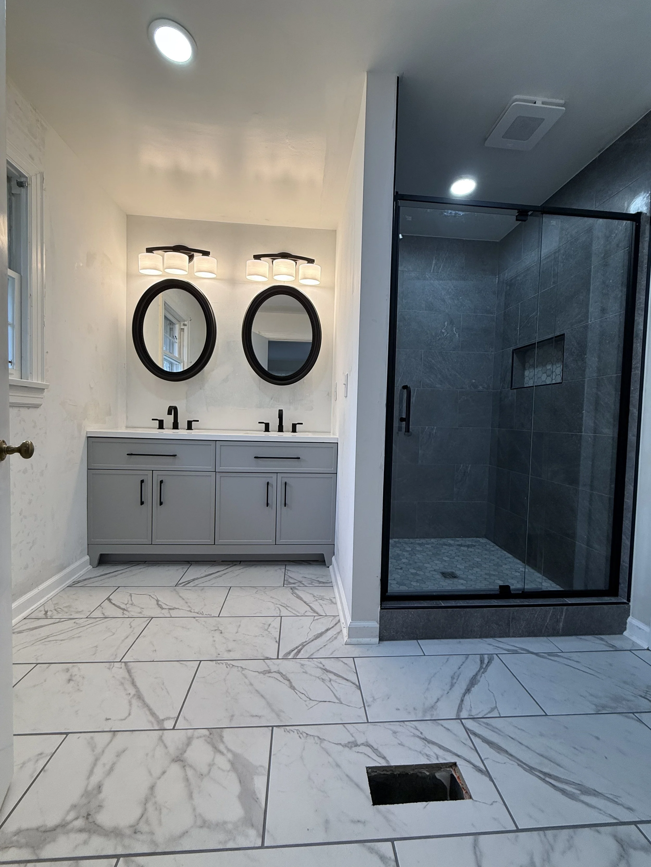 A modern bathroom featuring a double vanity with two mirrors and light fixtures, a glass-enclosed shower with dark gray tiles, and marble floor tiles. There is a small window and a hole in the floor, possibly for plumbing or wiring.