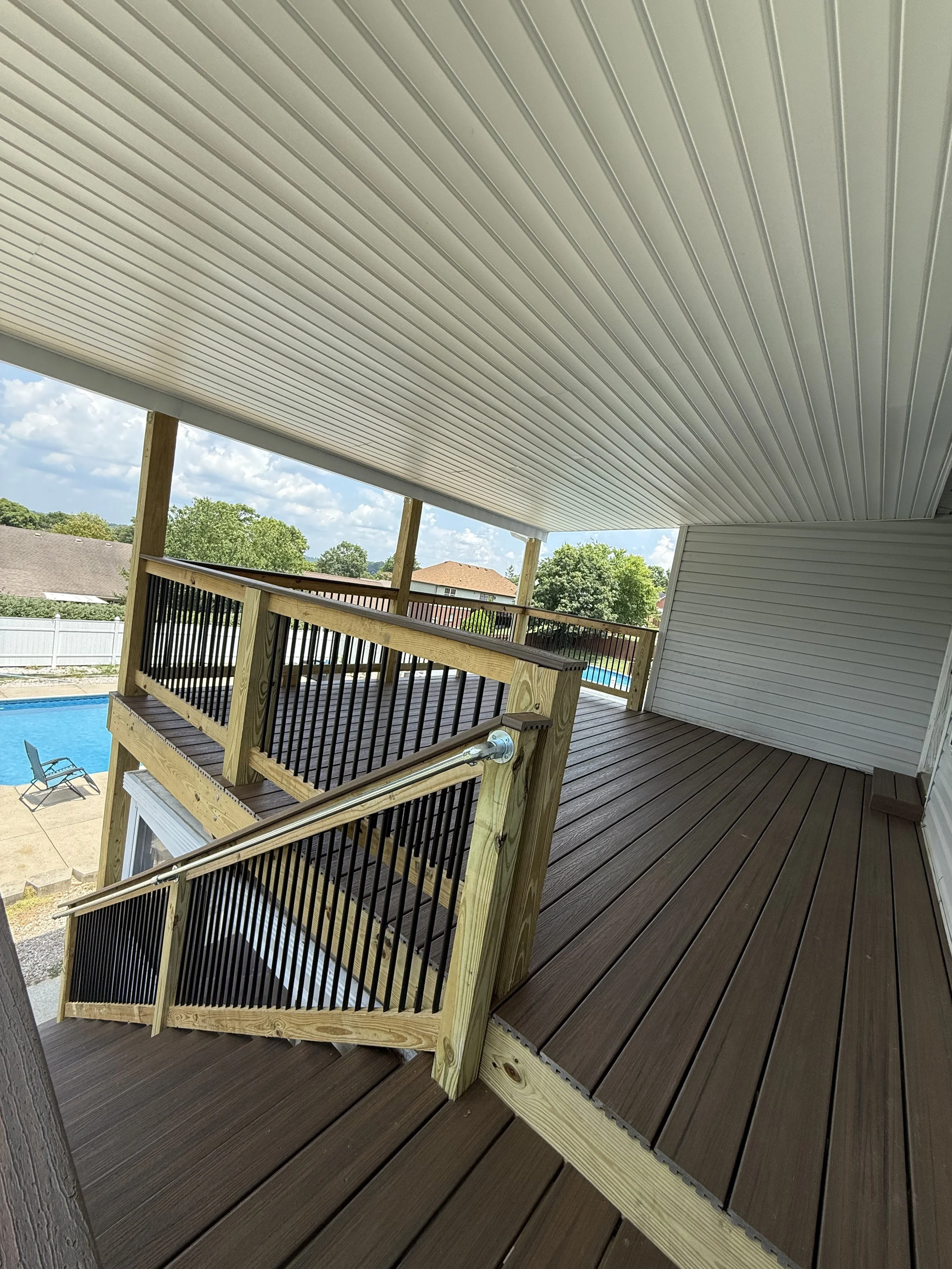 A two-story wooden balcony with black railing overlooking a backyard with a swimming pool, lounge chair, green trees, and a partly cloudy sky.