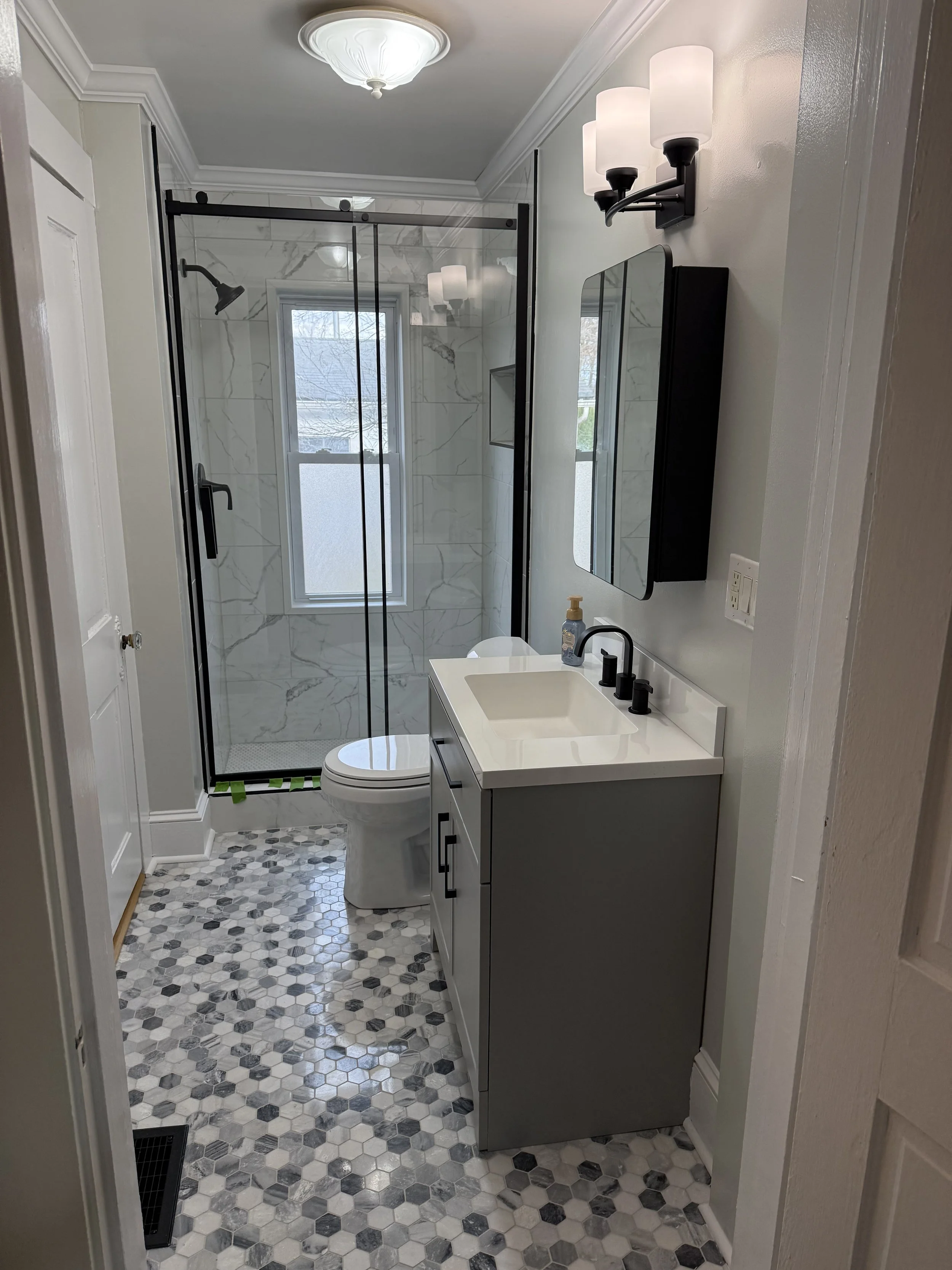 Modern small bathroom with a glass-enclosed shower, a white vanity with a sink, a mirror, and a light fixture with multiple bulbs. The bathroom has a hexagonal patterned floor in black, white, and gray tones.