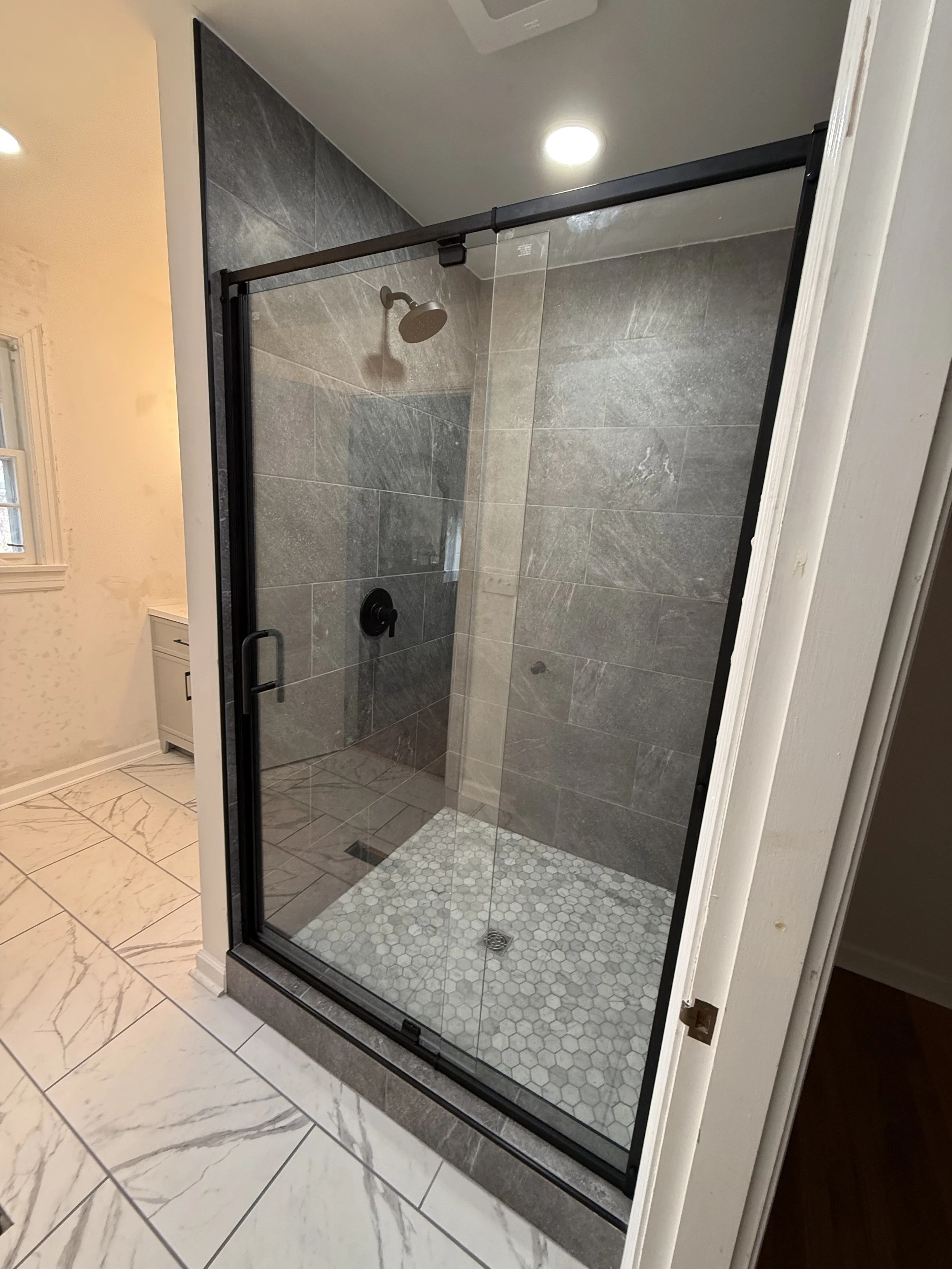 A modern walk-in shower with clear glass doors, gray tiled walls, a round showerhead, a black handle, and a white hexagonal tiled shower floor.