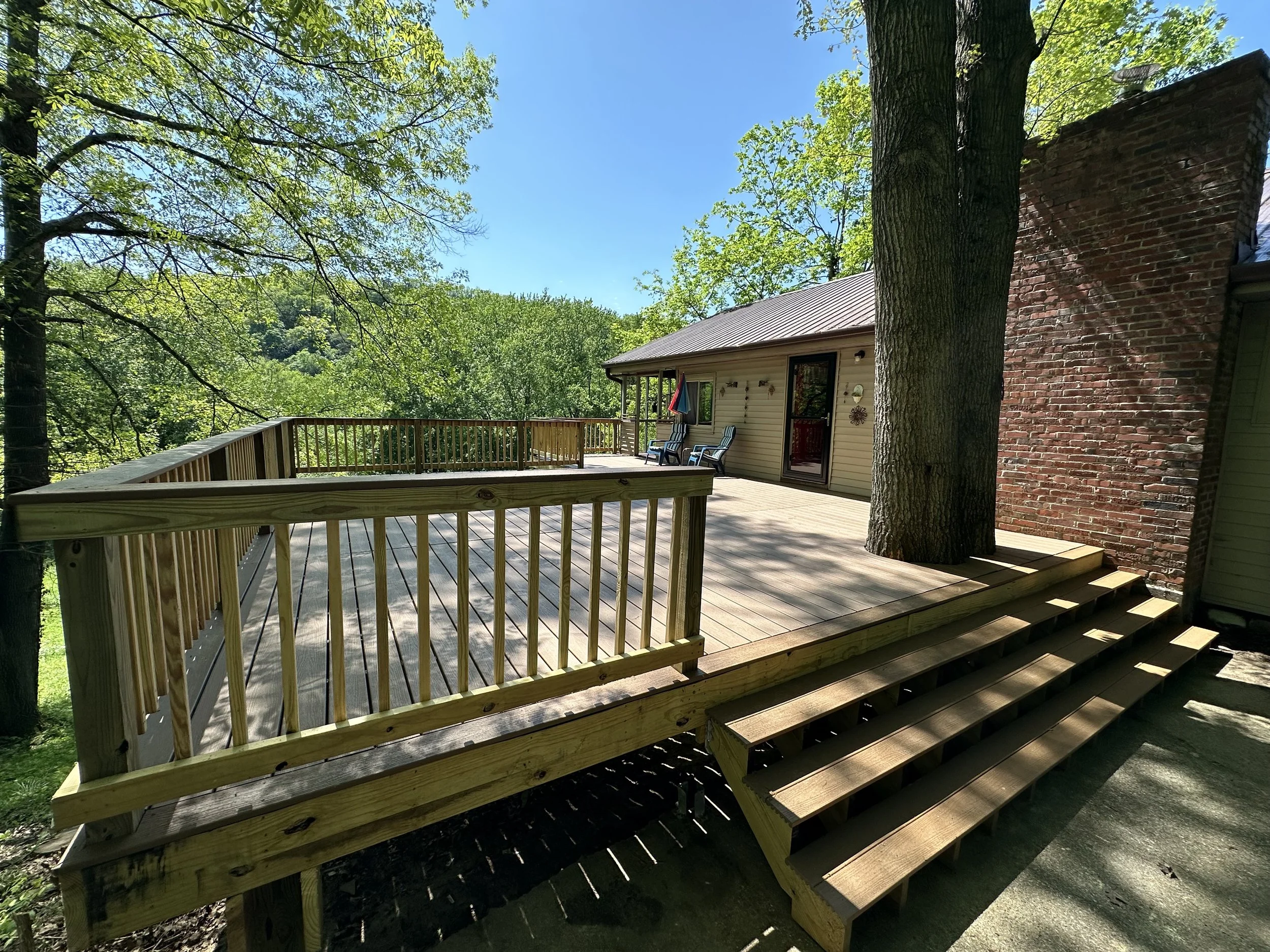 Wooden deck with stairs, attached to a house, surrounded by trees and greenery, on a sunny day.