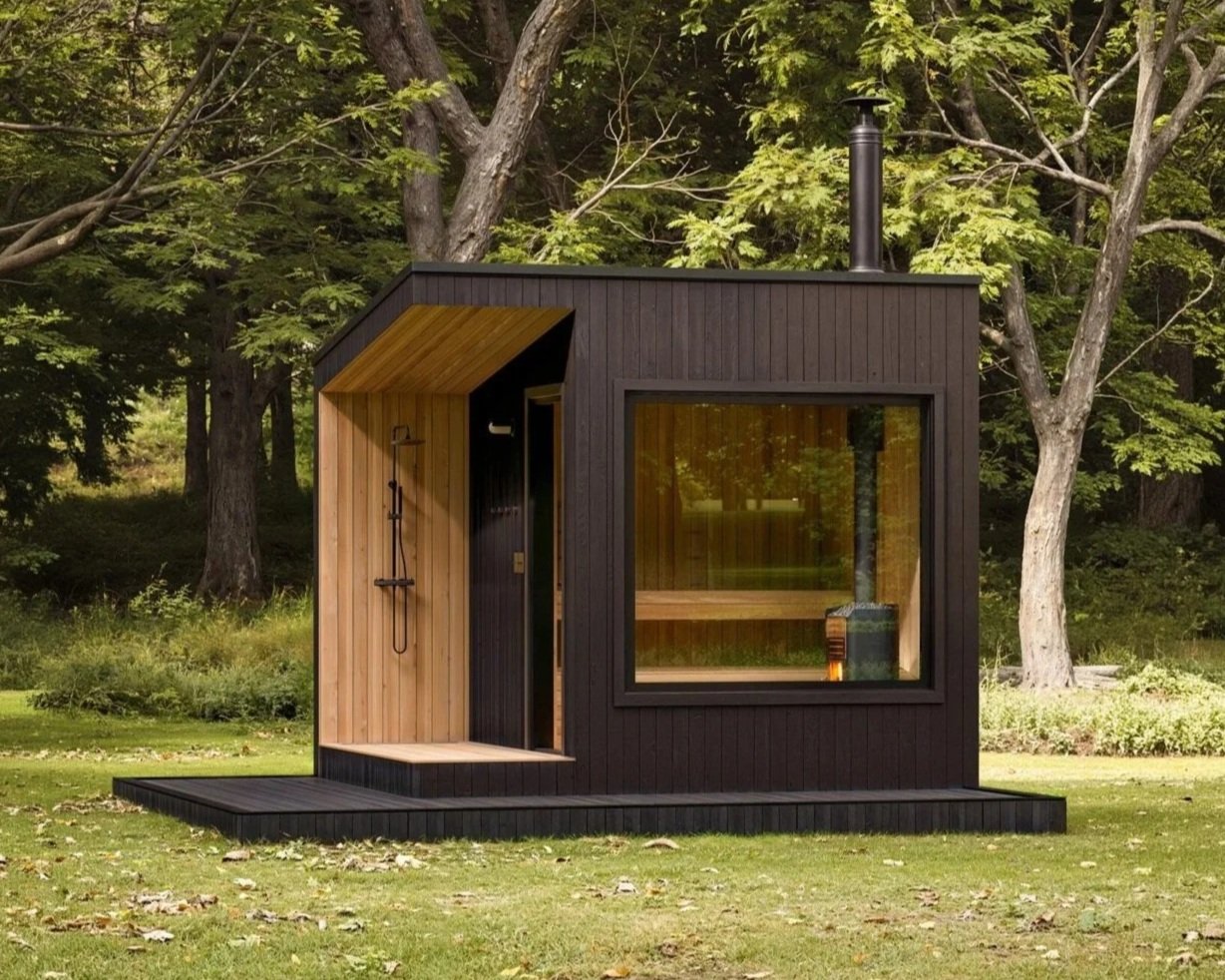 Small modern black house with large window, wood accents, outdoor shower, and chimney, situated in a forest clearing.