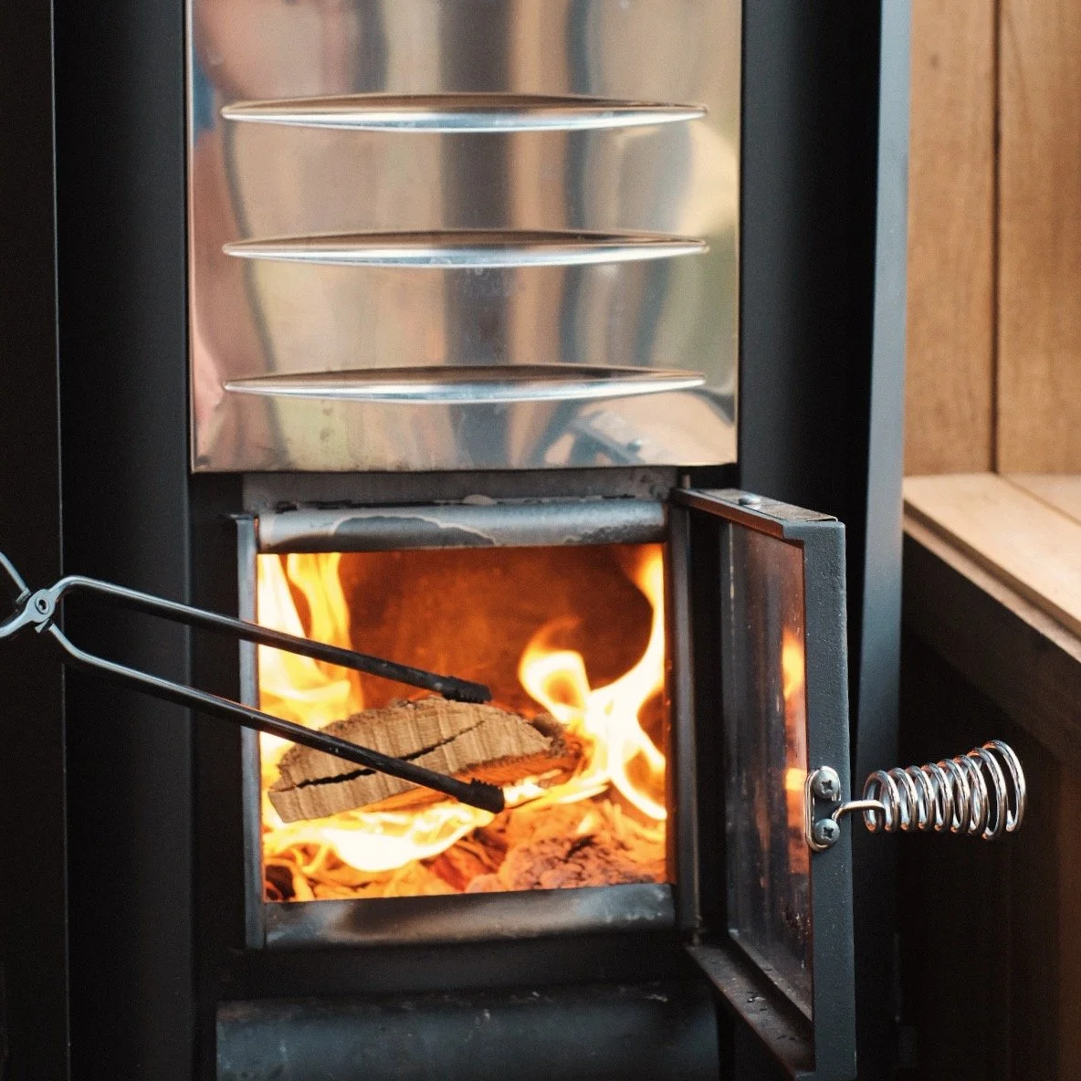 A small wood-burning stove with a metal door open, showing flames and burning firewood inside, with metal shelves above.