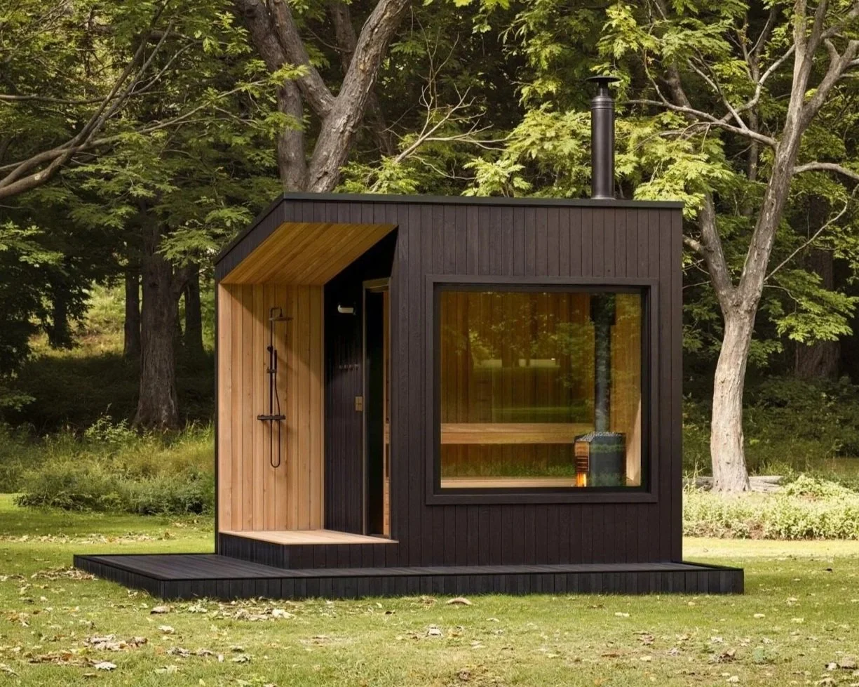 Small modern black and wood cabin with large window and outdoor shower, set in a forest clearing.