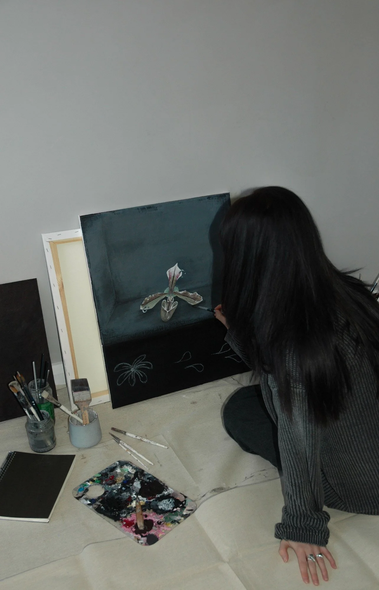 Art student painting a realistic image of a flower on a box-shaped canvas in a studio. The artist has supplies like brushes, a palette, and a notebook nearby.