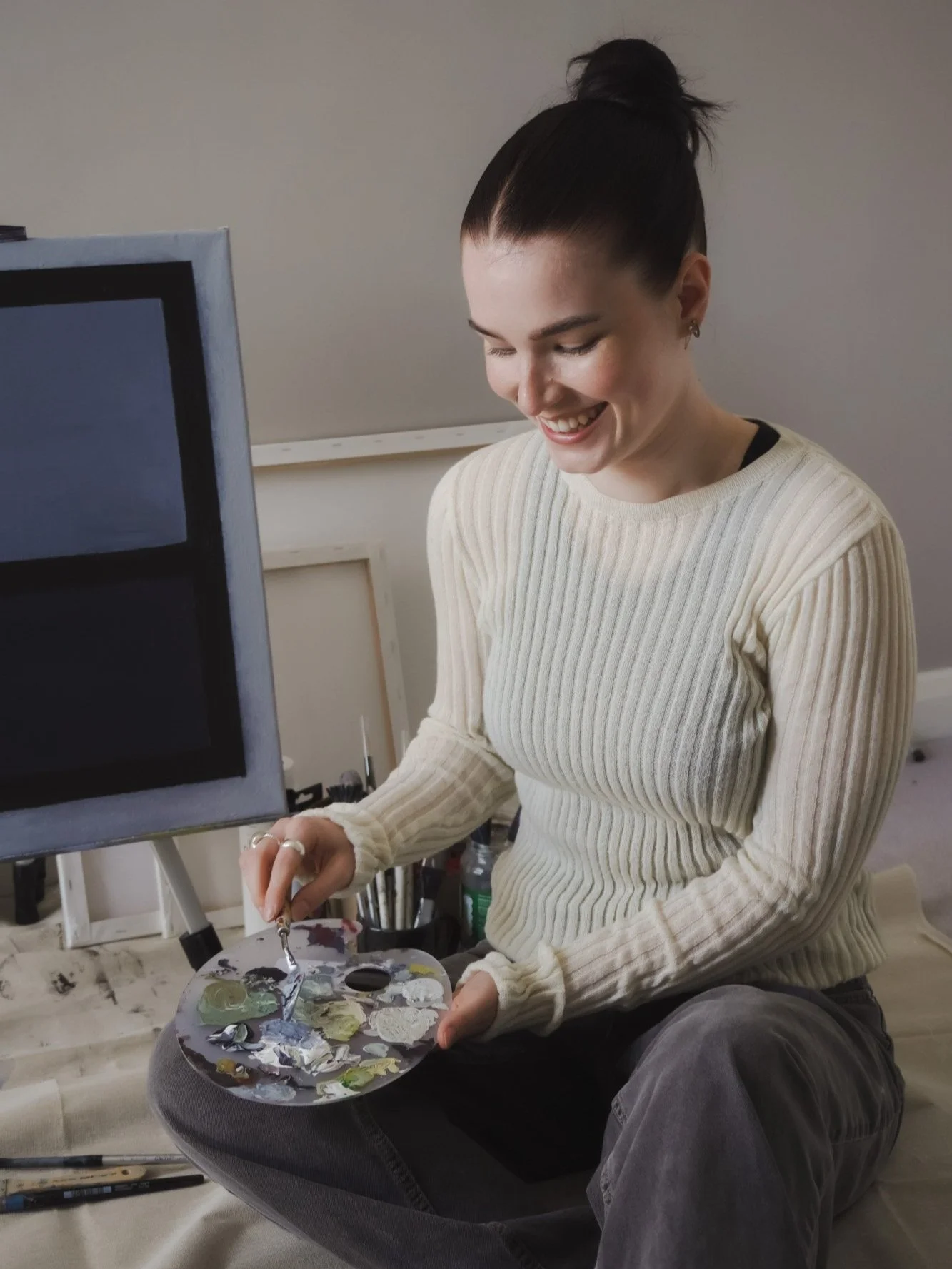 A woman with dark hair in a bun, wearing a cream-colored ribbed sweater, smiling while sitting and mixing paint on a palette during an art session.