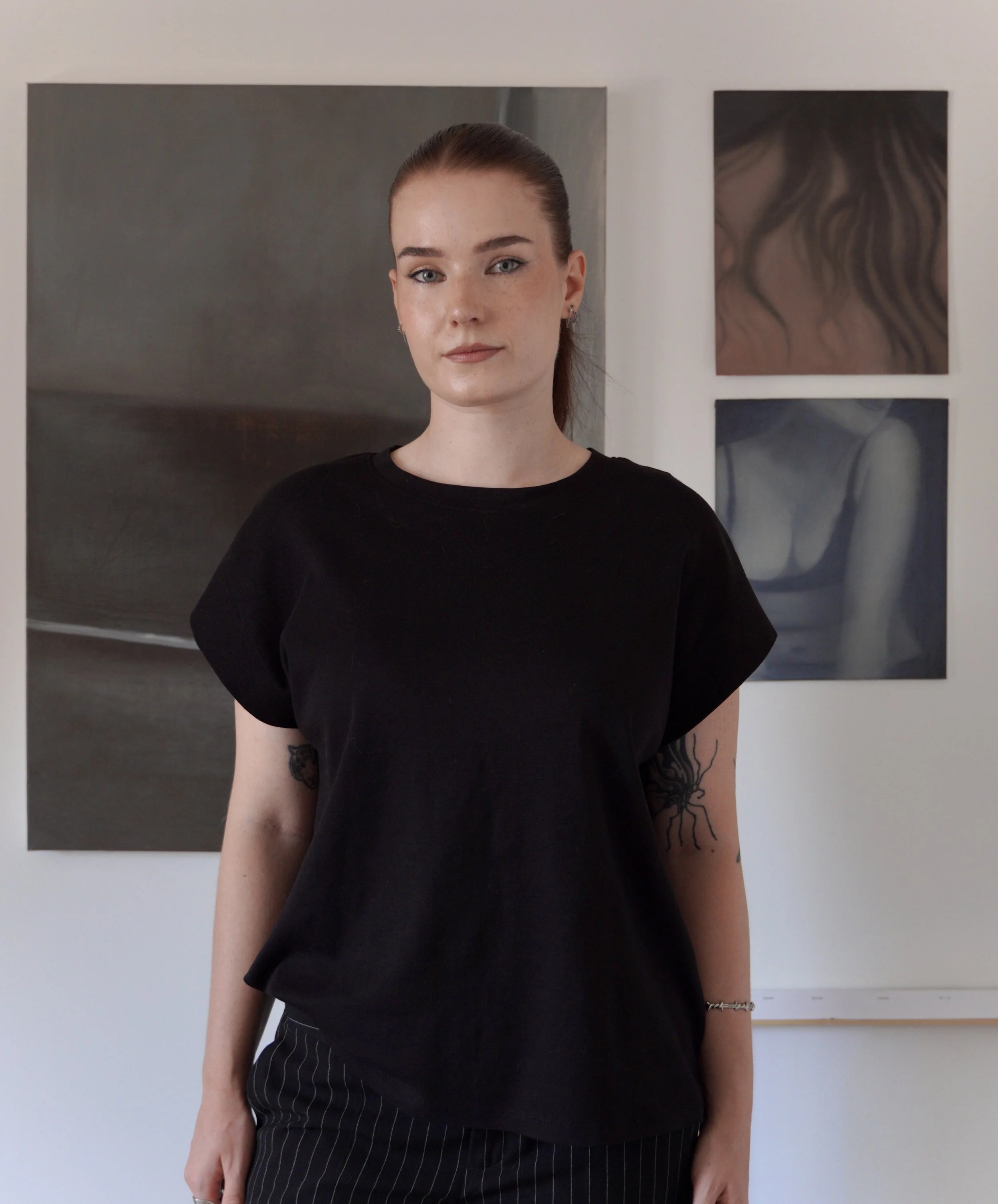 A woman with fair skin, freckles, and reddish-brown hair pulled back, wearing a black shirt and black pinstripe pants, standing in front of a white wall with artwork.