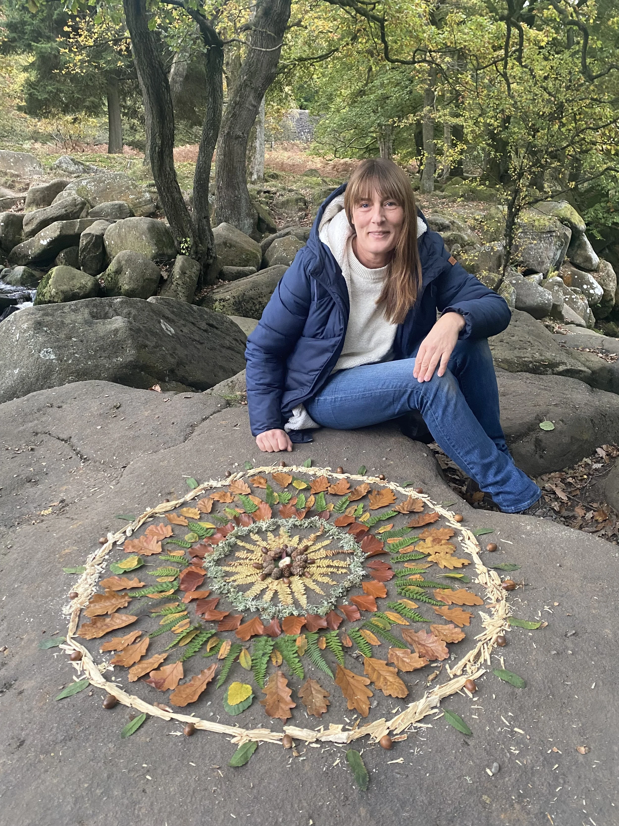 A woman in a blue jacket and jeans sitting on a large rock in a wooded area with a mandala made of leaves, pinecones, and other natural materials in front of her.
