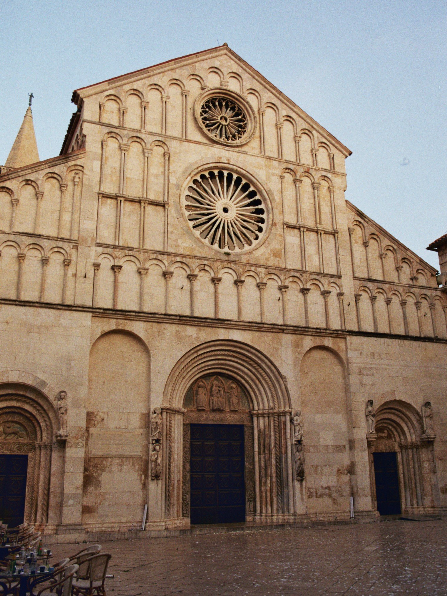 Fassade einer historischen gotischen Kirche mit großen Toren undrosafarbenen Steinwänden, zwei großen Rosettenfenstern und einem steinernen Feinsaunar mit Skulpturen über dem Eingang.