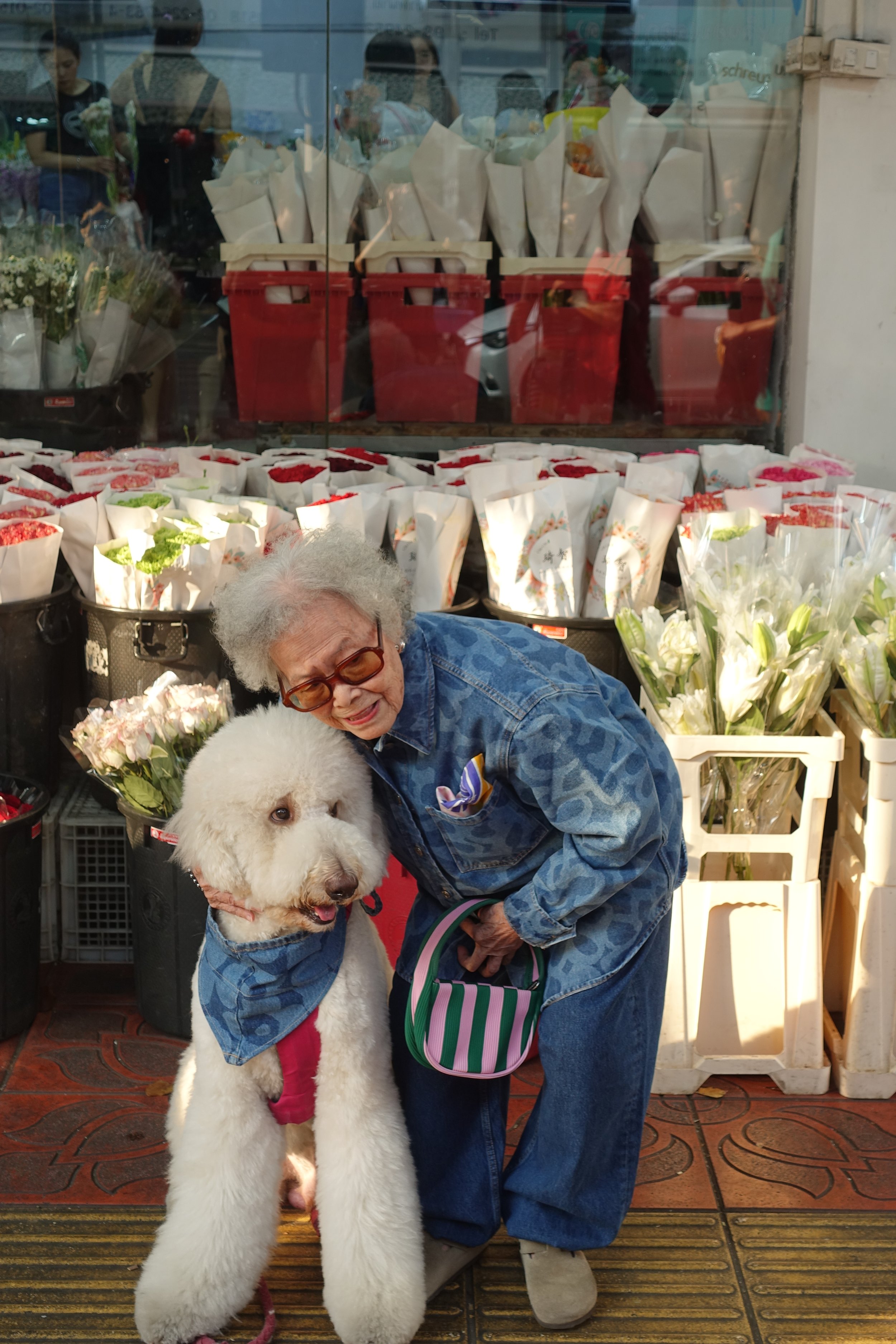 Ältere Frau mit Brille und weißem Zopf umarmt großen weißen Hund vor Blumenladen.