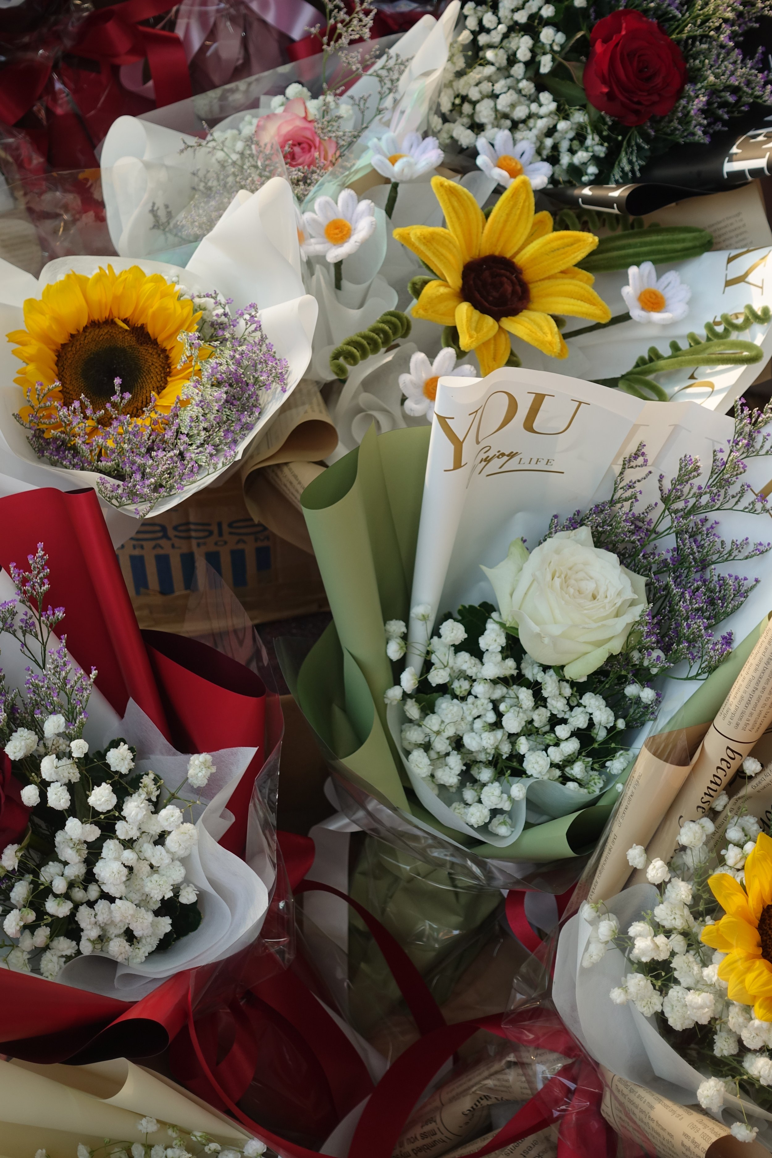 Verschiedene Blumensträuße mit Sonnenblumen, Rosen, Gänseblümchen und Schleierkraut, eingewickelt in farbiges Papier und Folie.