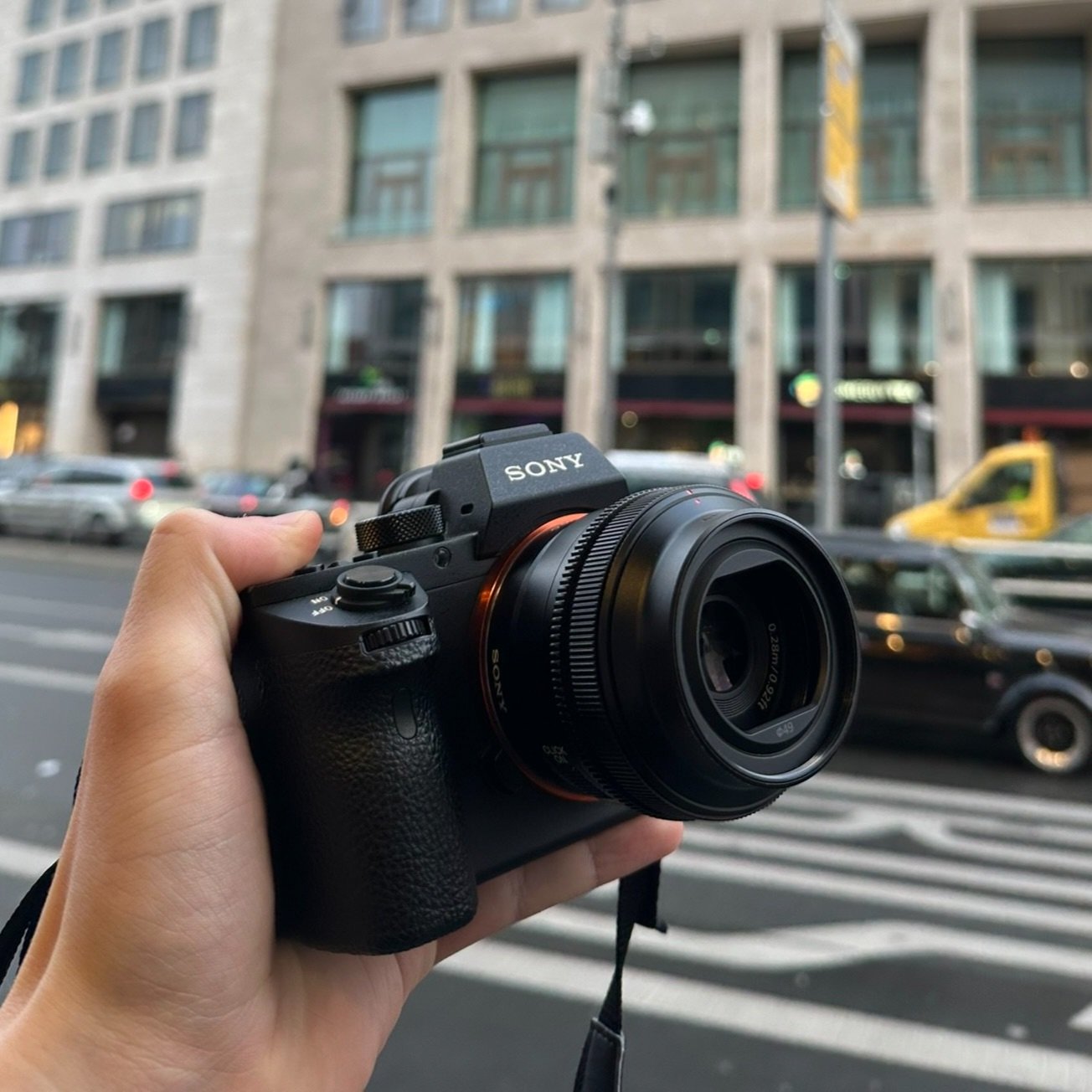 Hand hält eine Sony Kamera vor der Straße, im Hintergrund sind Gebäude, Autos und eine Fußgängerampel sichtbar.