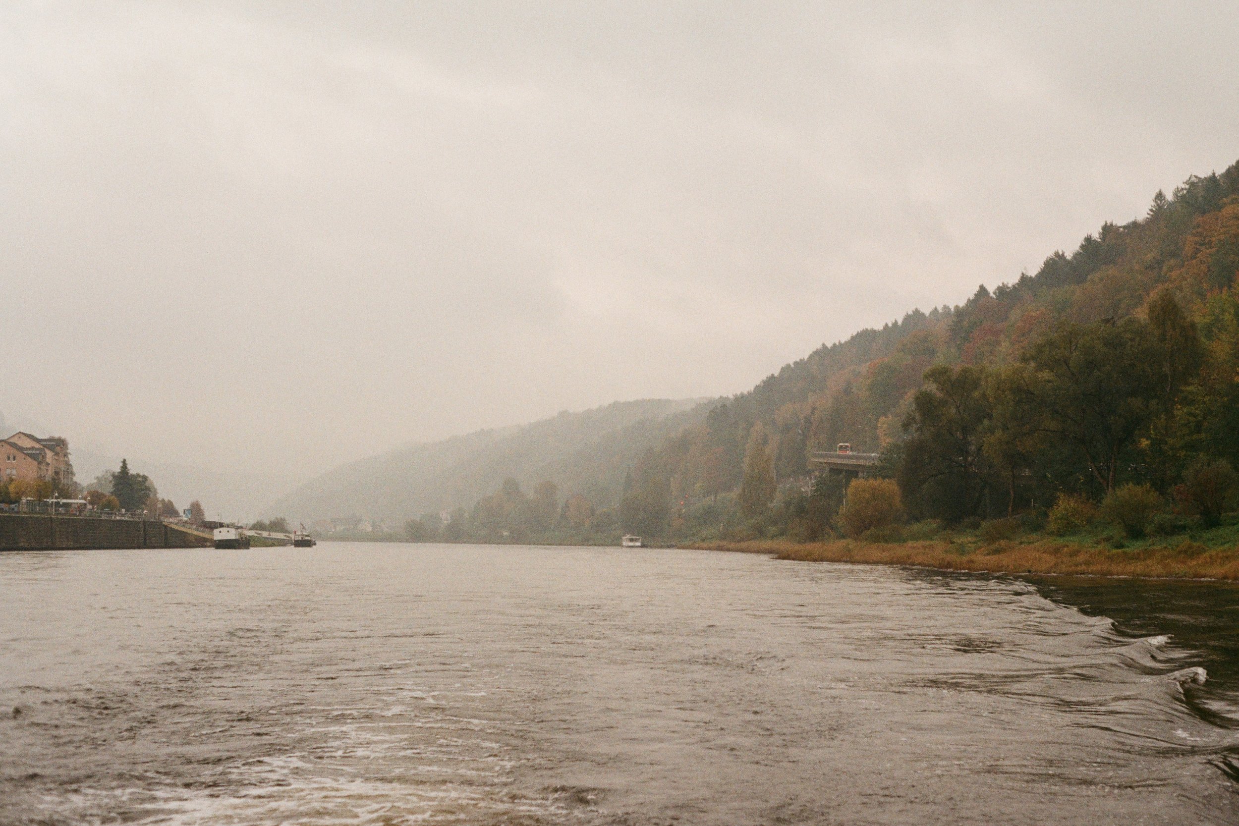 zwei Boote auf einem Fluss, umgeben von bewaldeten Hängen und Gebäuden am Ufer, an einem nebligen Tag