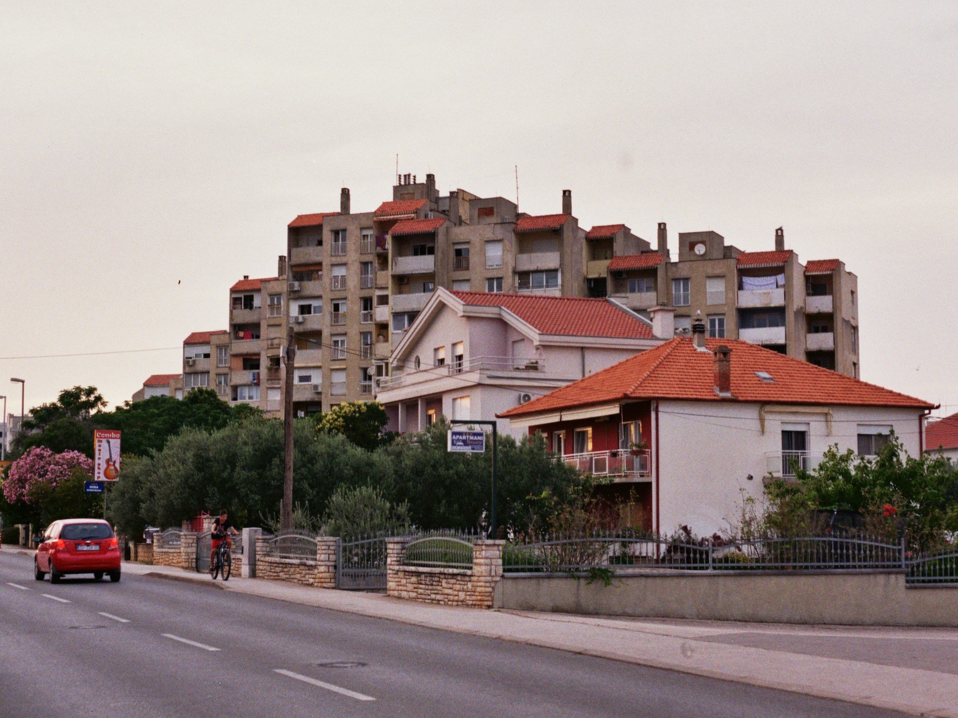 Stadthäuser und ein Hochhaus in einer Wohnsiedlung, mit Autos auf der Straße und Bäumen im Vordergrund, bei Sonnenuntergang