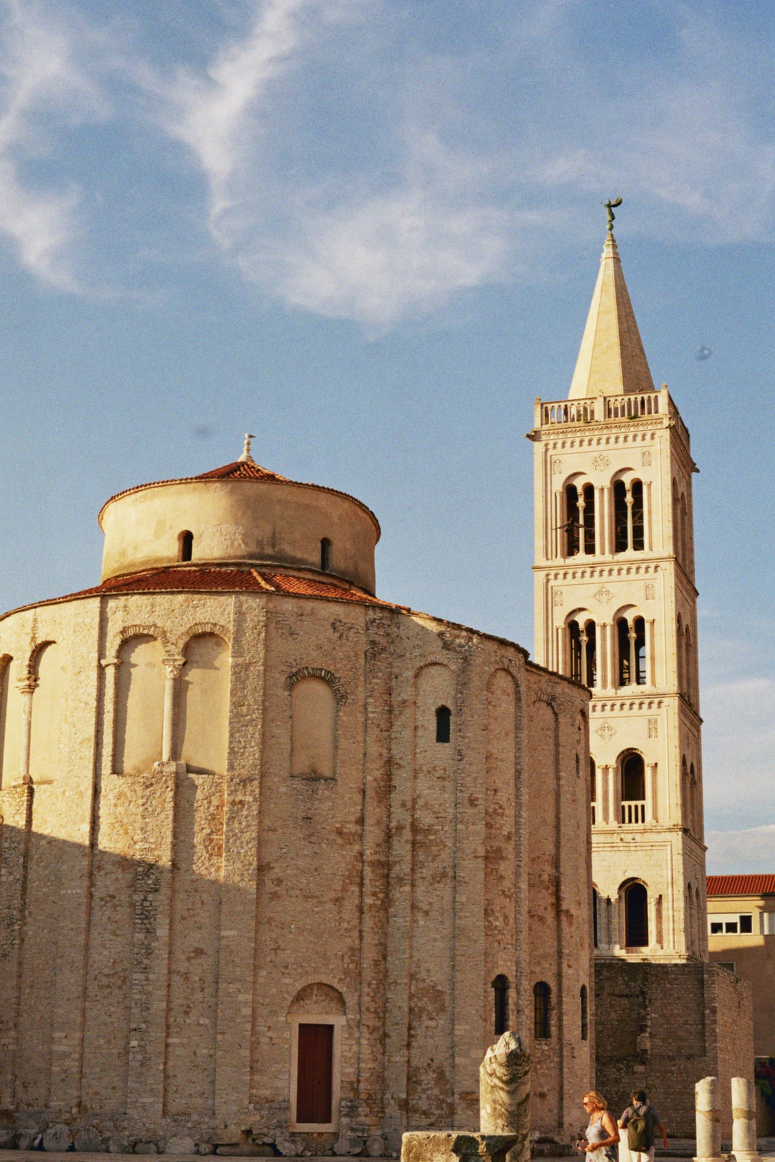 Historische Kirche mit runder Bauform und hohem Turm im Sonnenlicht, im Vordergrund Personen und alte Säulen.