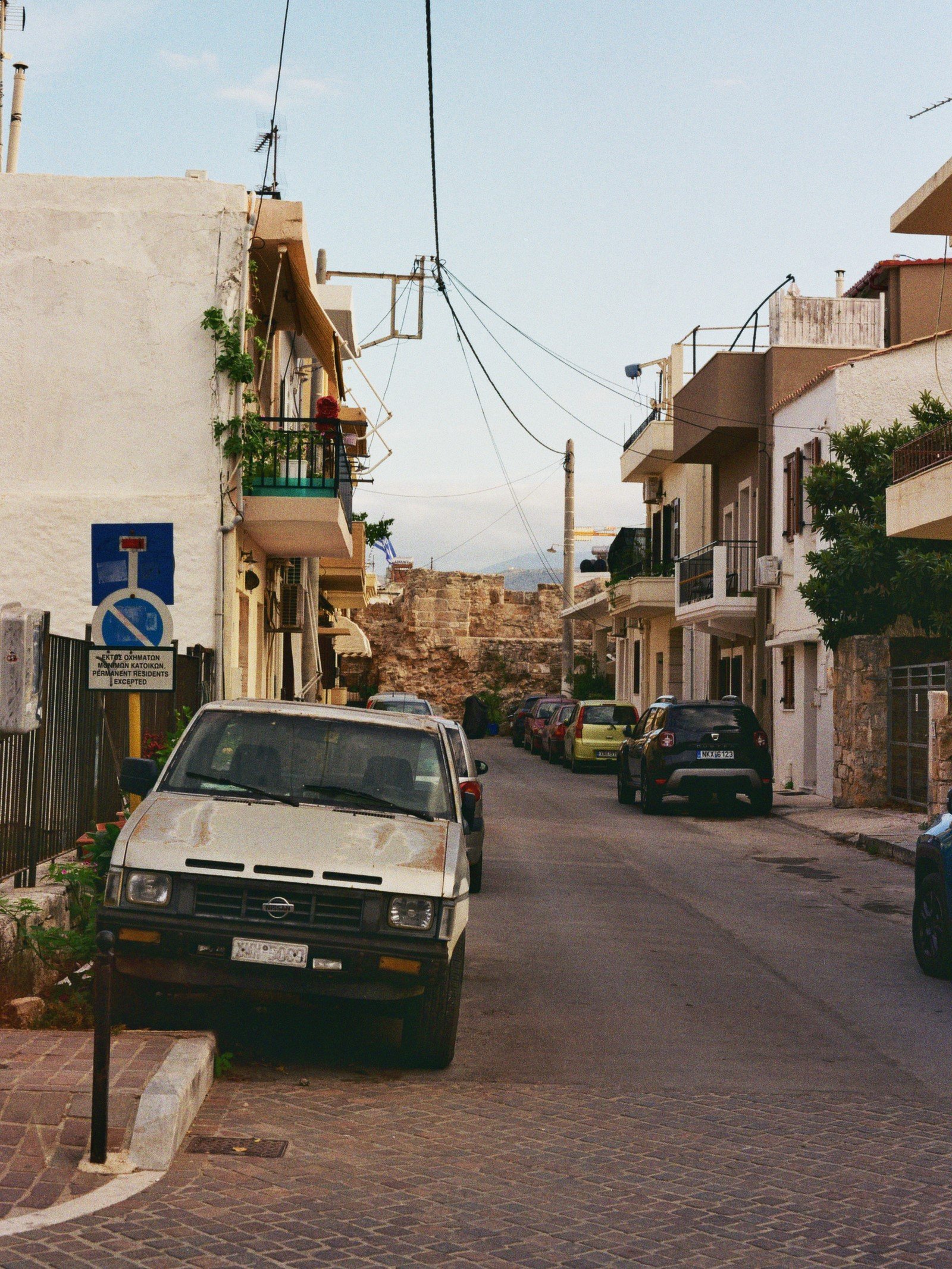 Straße in einer Stadt mit parkenden Autos und Mehrstöckigen Wohnhäusern auf beiden Seiten. Es gibt eine Wand im Hintergrund und eine Stromleitung unter dem blauen Himmel.