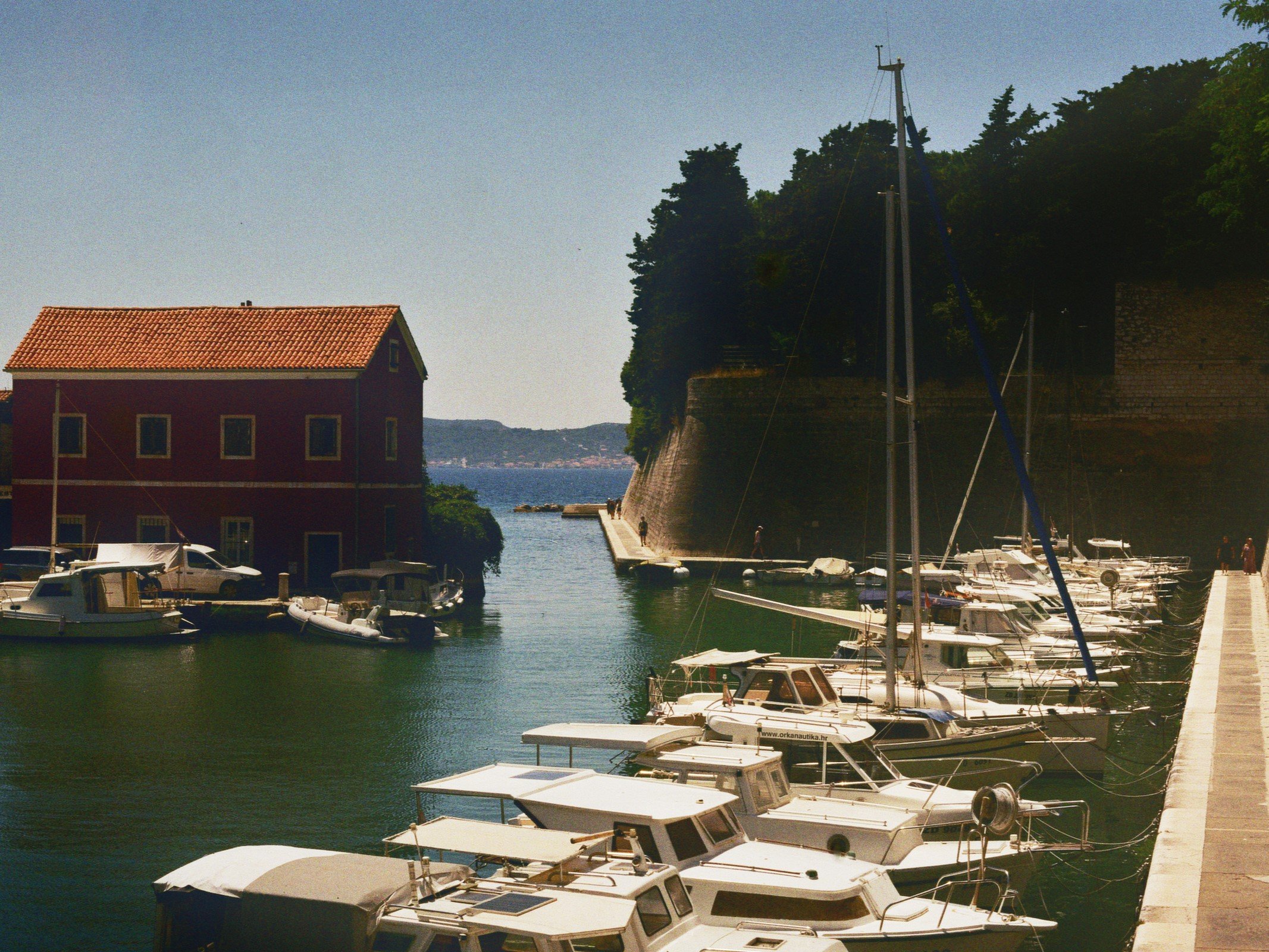 Boots im Hafen neben einem roten Haus mit rotem Dach, im Hintergrund Wasser und Bäume.