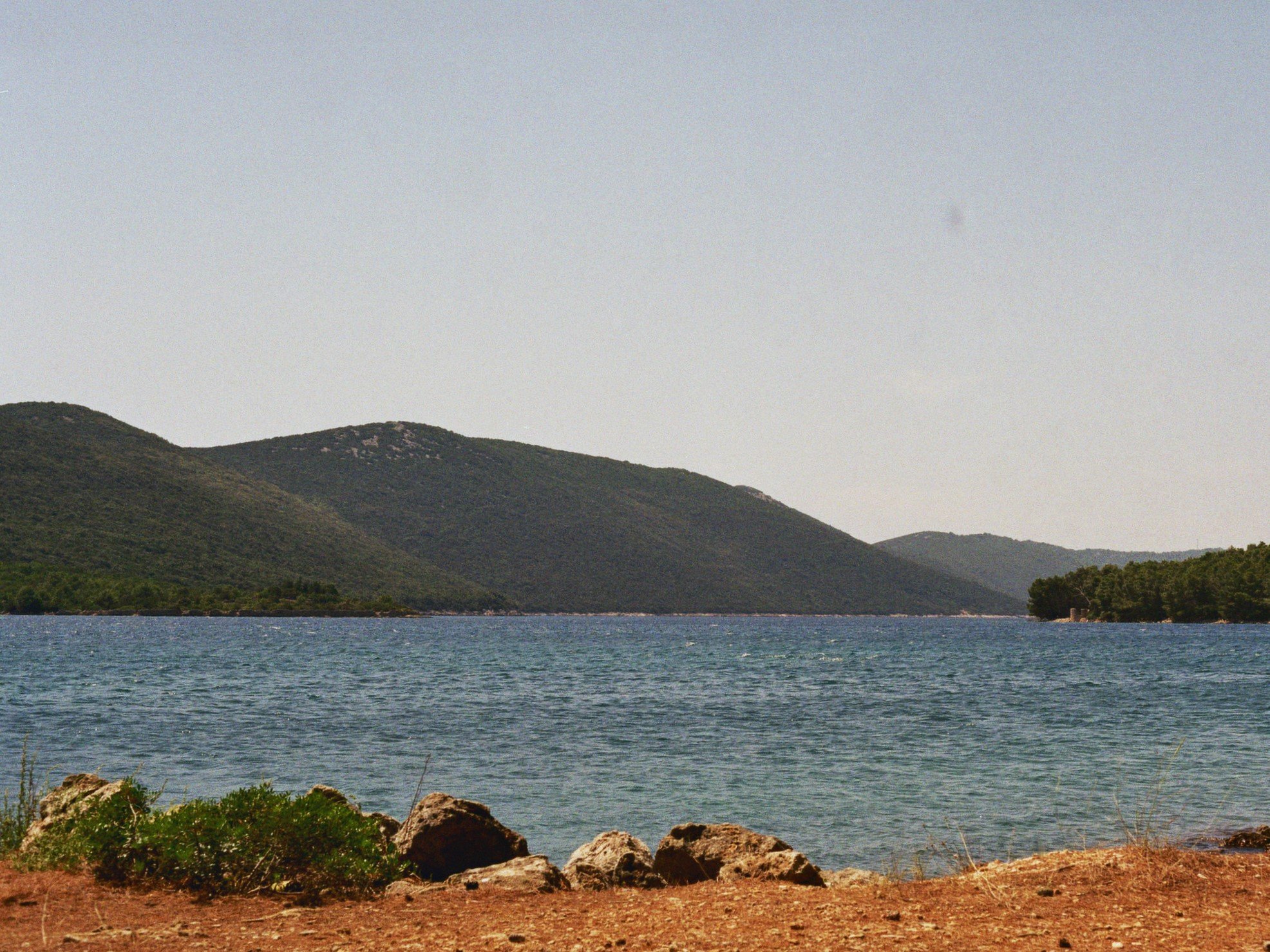 Blick auf einen See mit Bergen im Hintergrund, felsige Uferkante im Vordergrund, leicht bewölkter Himmel.