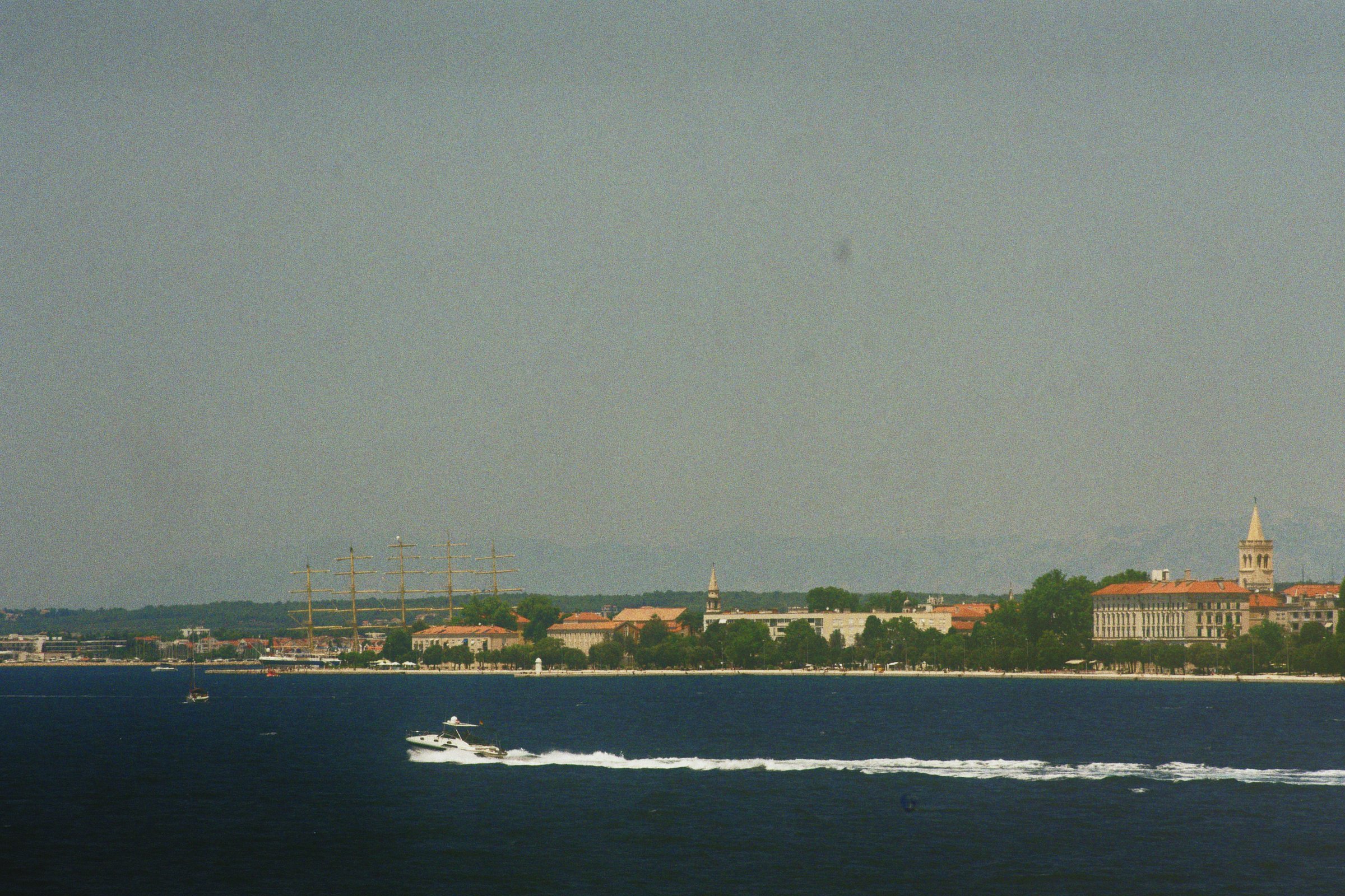 Blick auf eine Stadt am Wasser mit Segelschiff, Motorboot und historischem Gebäudekomplex, inklusive Kirchtürmen, bei Sonnenuntergang.