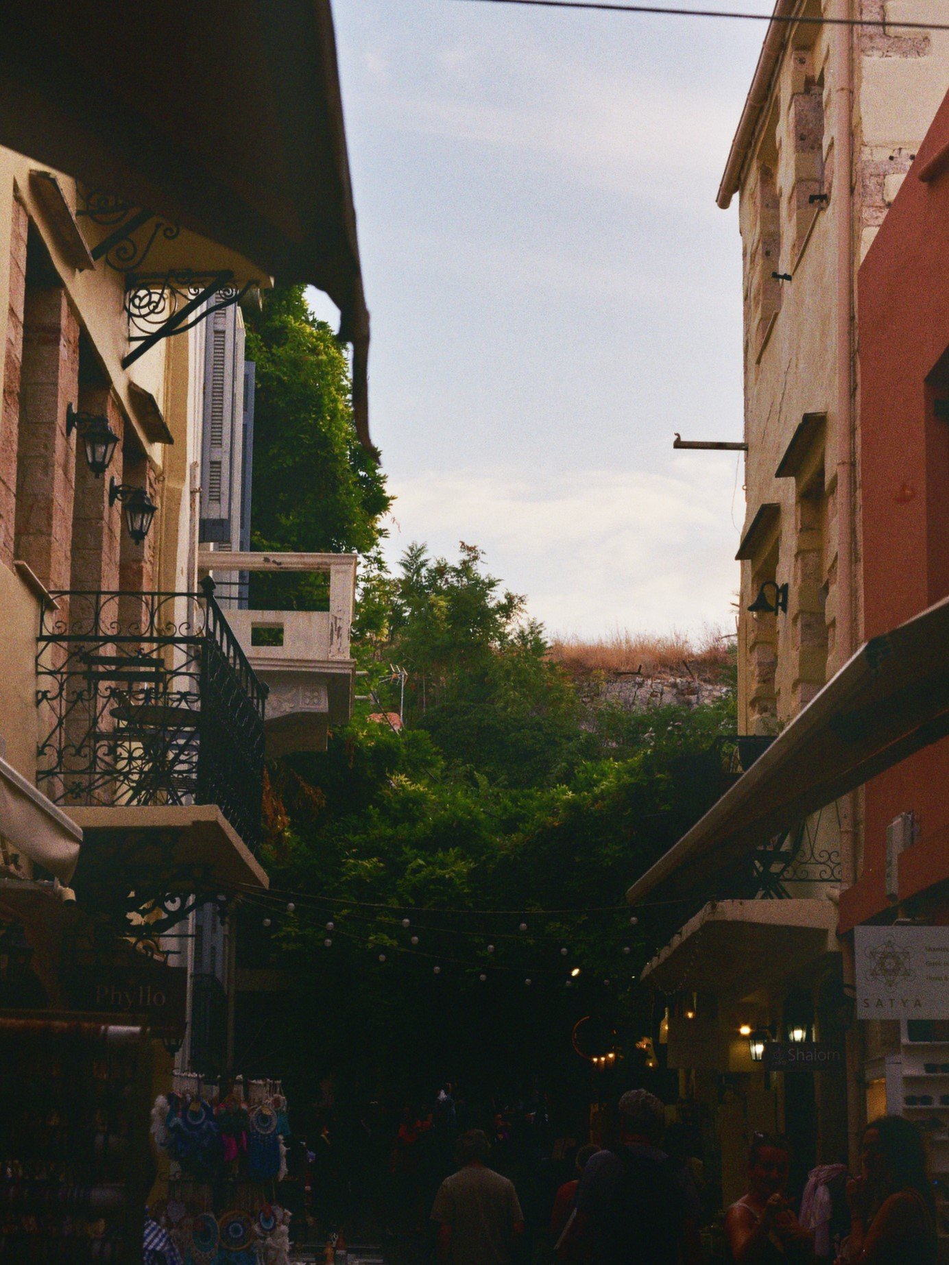 Ein belebter Marktplatz in einer Stadt mit mehreren Geschäften und Menschen, umgeben von Gebäuden mit Balkonen und grünen Bäumen im Hintergrund.