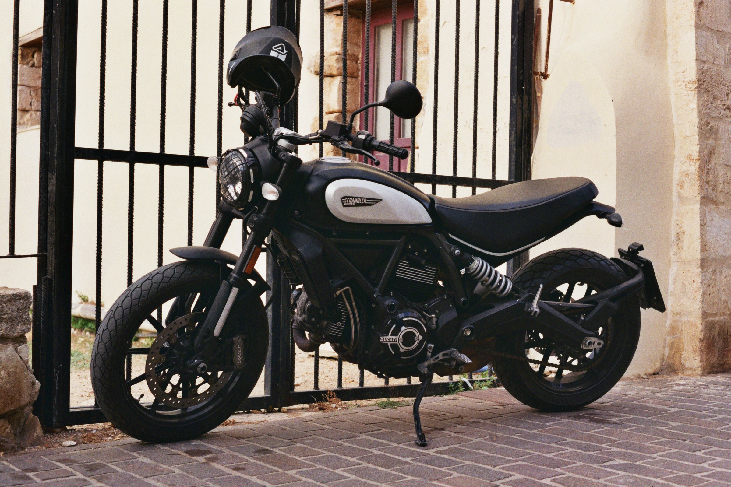 Ein schwarzes Motorrad mit weiß-hellgrauen Akzenten, parkiert auf gepflastertem Boden vor einem Metalltor.