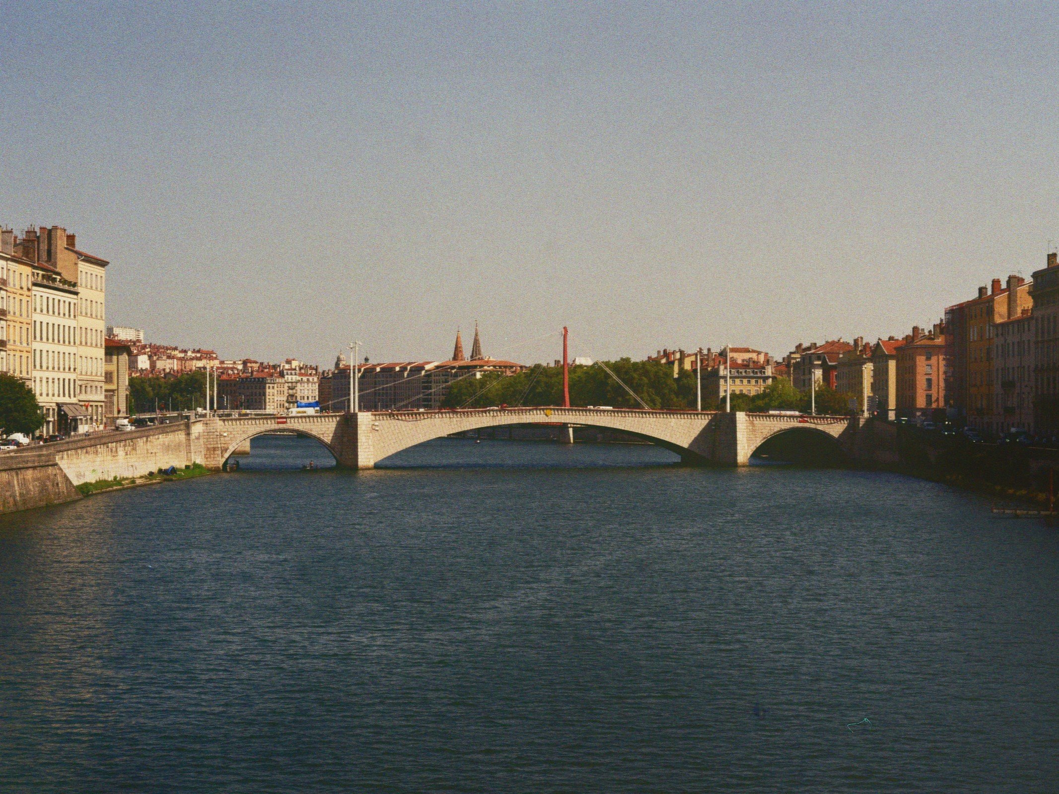 Ein Fluss mit einer Steinbrücke darüber, umgeben von historischen Gebäuden in einer Stadt, unter klarem Himmel.