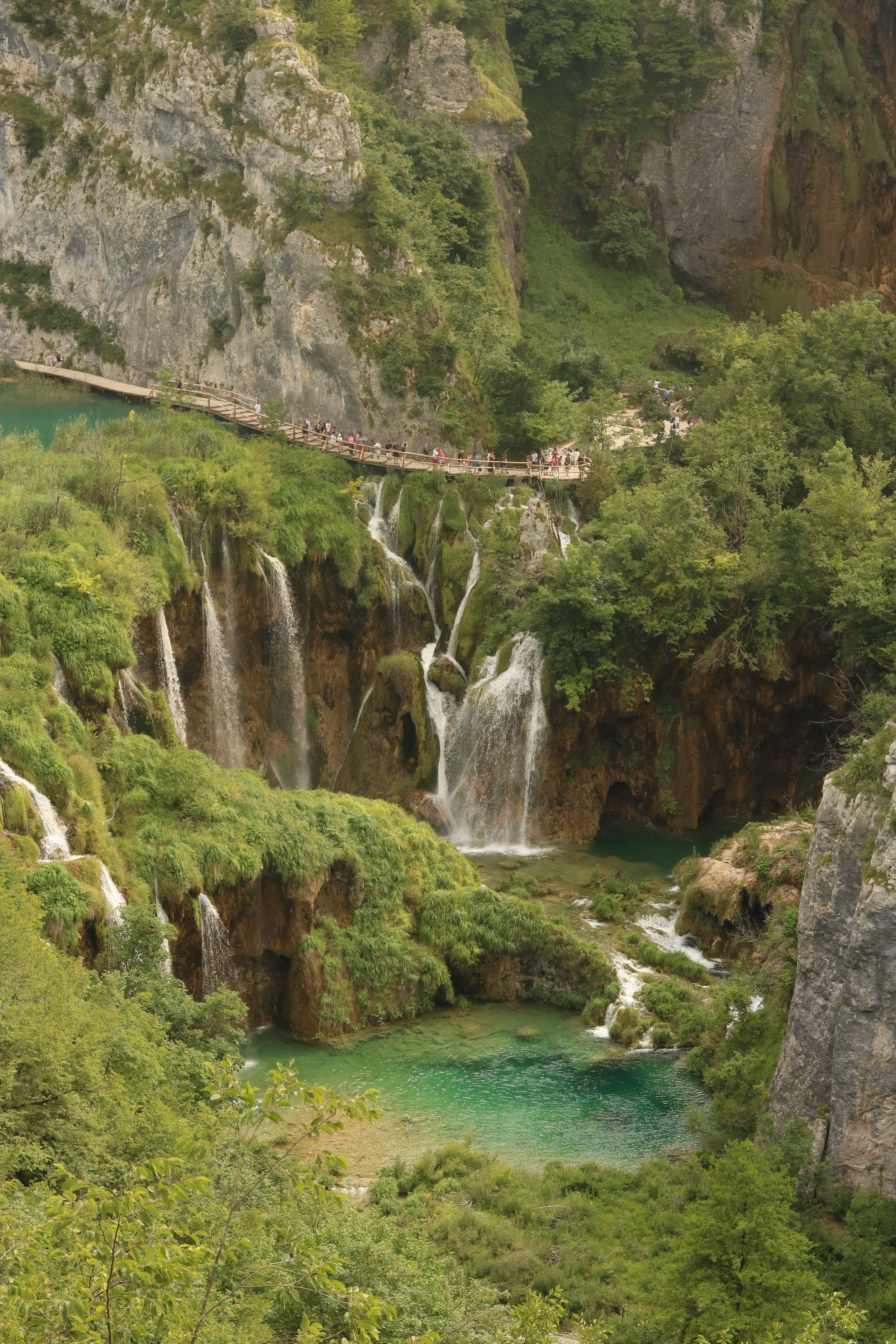 Sicht auf einen Wasserfall in einer grünen, felsigen Schlucht, mit Holzsteg und Besuchern.