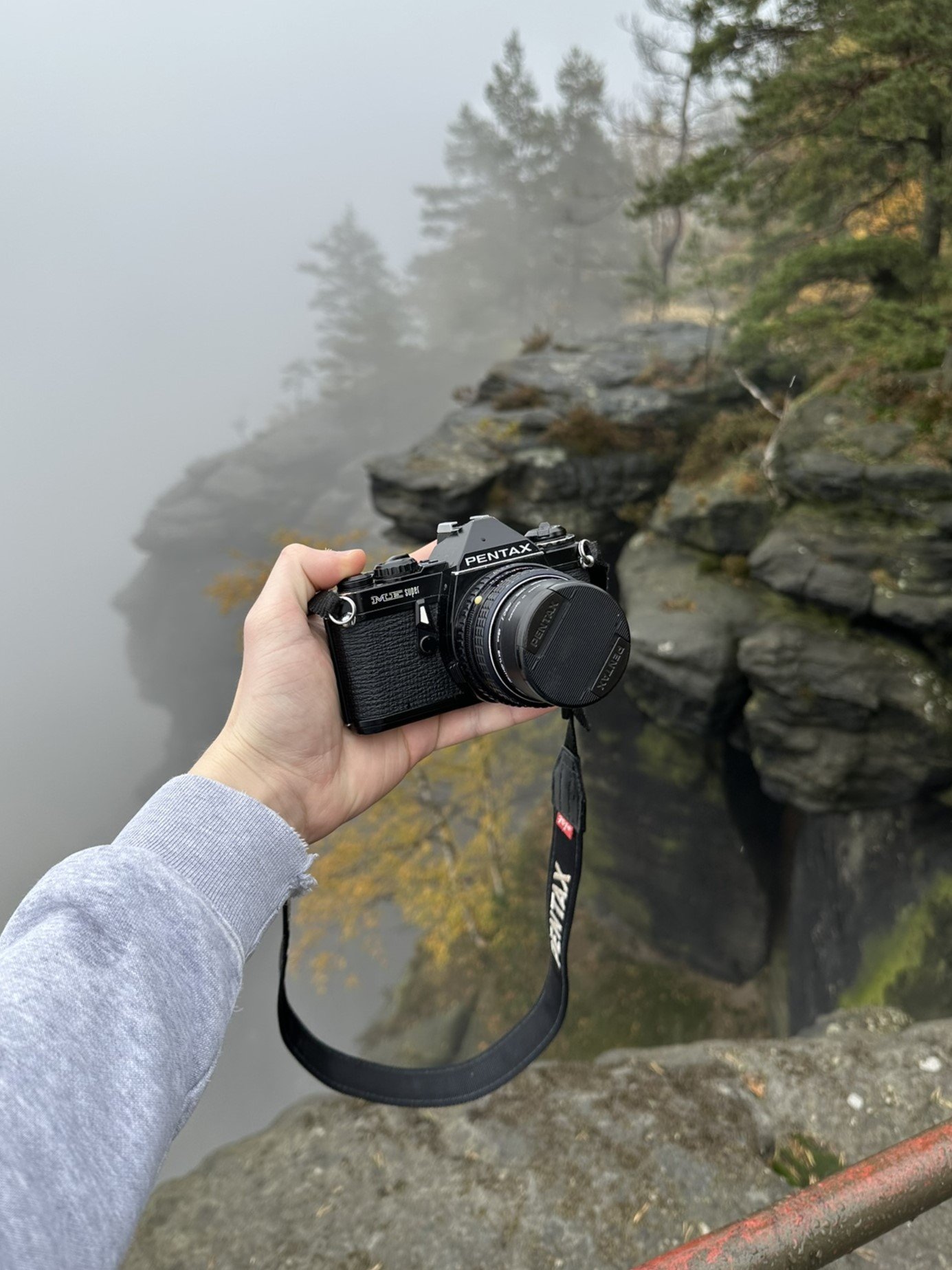 Hand hält eine Pentax Kamera vor nebeliger Flusslandschaft mit Felsen und Bäumen.