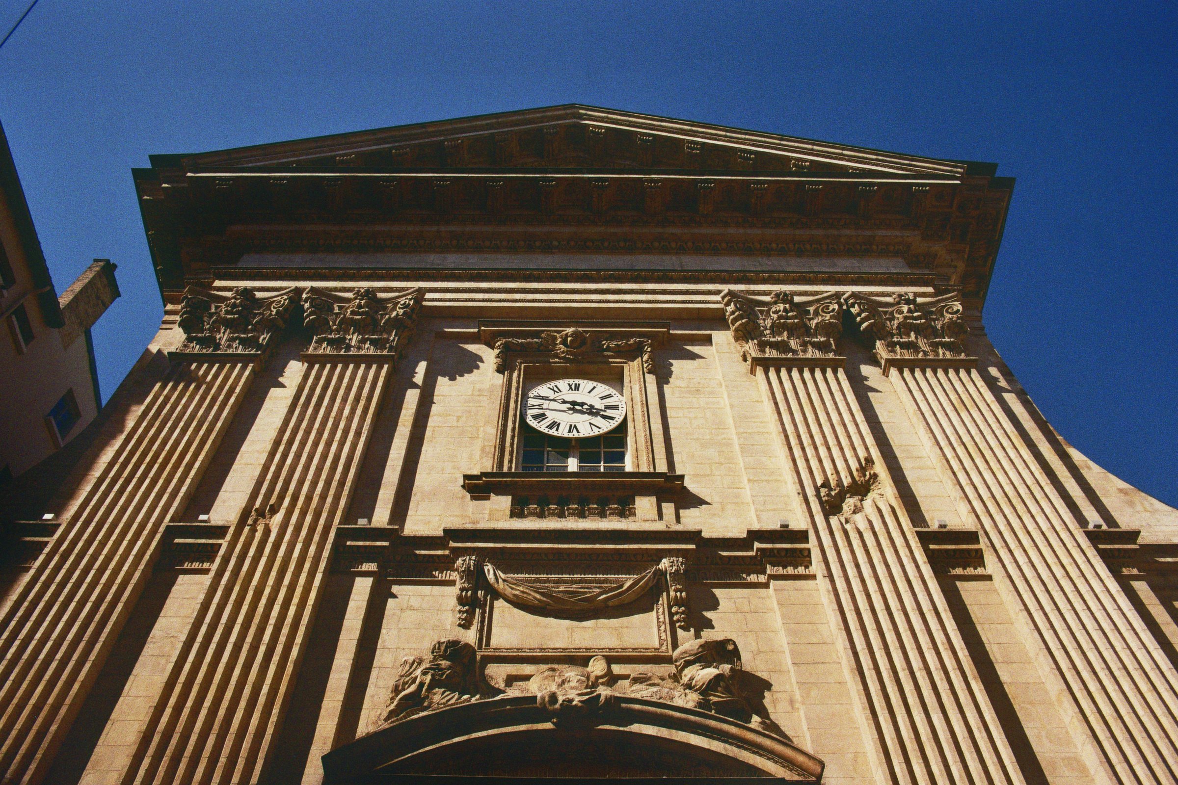 Ansicht eines historischen Gebäudes mit Uhr, zu beiden Seiten Säulen und Verzierungen, bei klarem Himmel.
