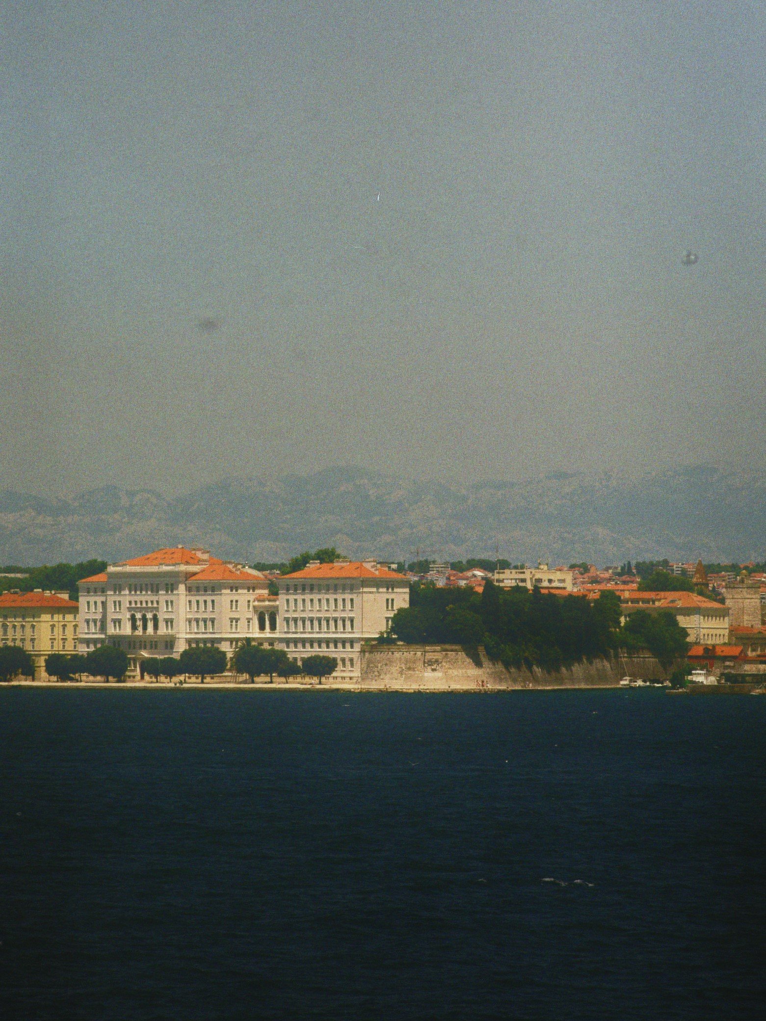 Blick auf eine Küstenstadt mit historischen Gebäuden, Wasser im Vordergrund und Berge im Hintergrund.
