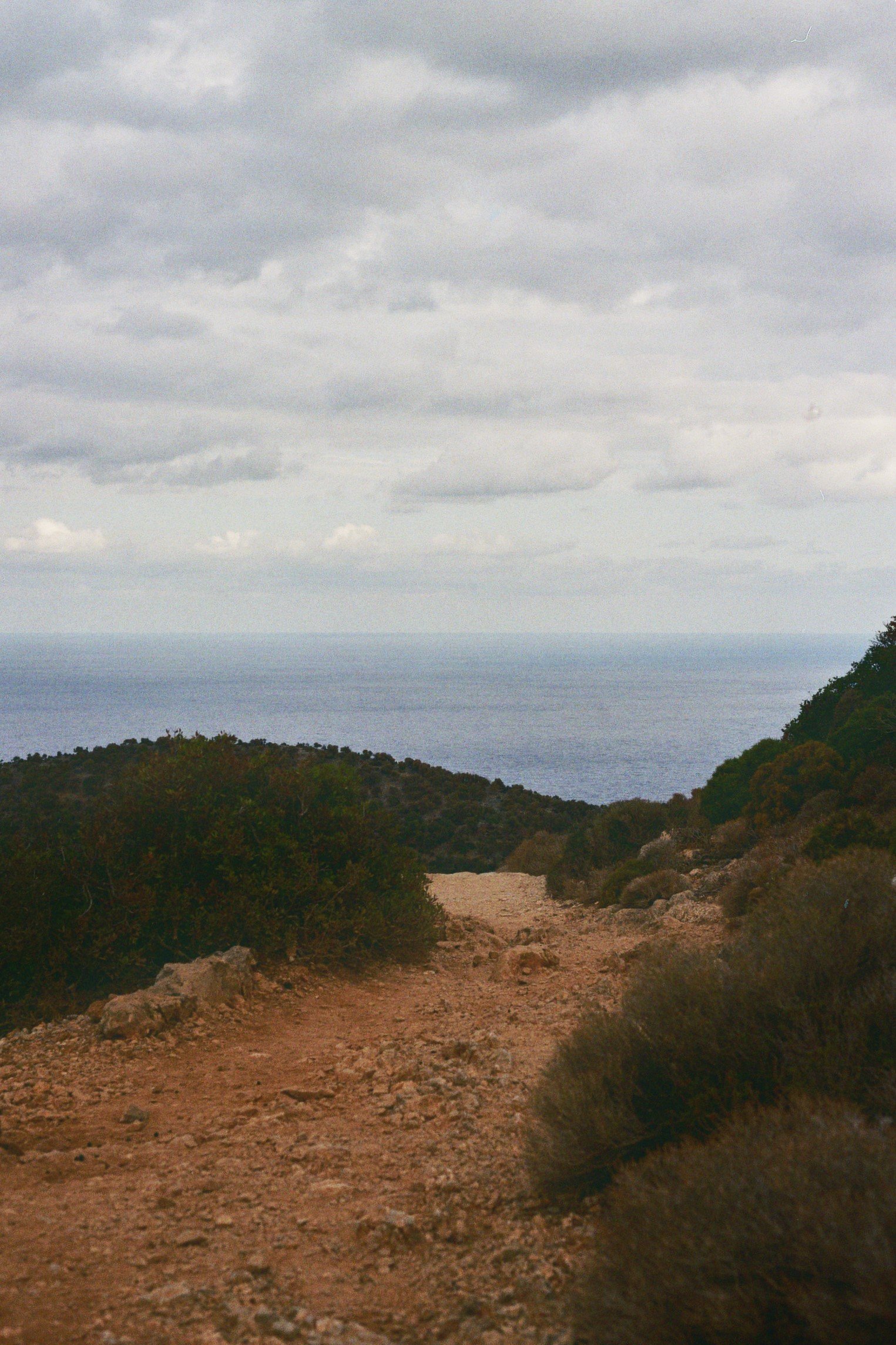 Ein unbefestigter Wanderweg führt durch eine trockene, buschige Landschaft, mit Blick auf das Meer und bewölkten Himmel im Hintergrund.