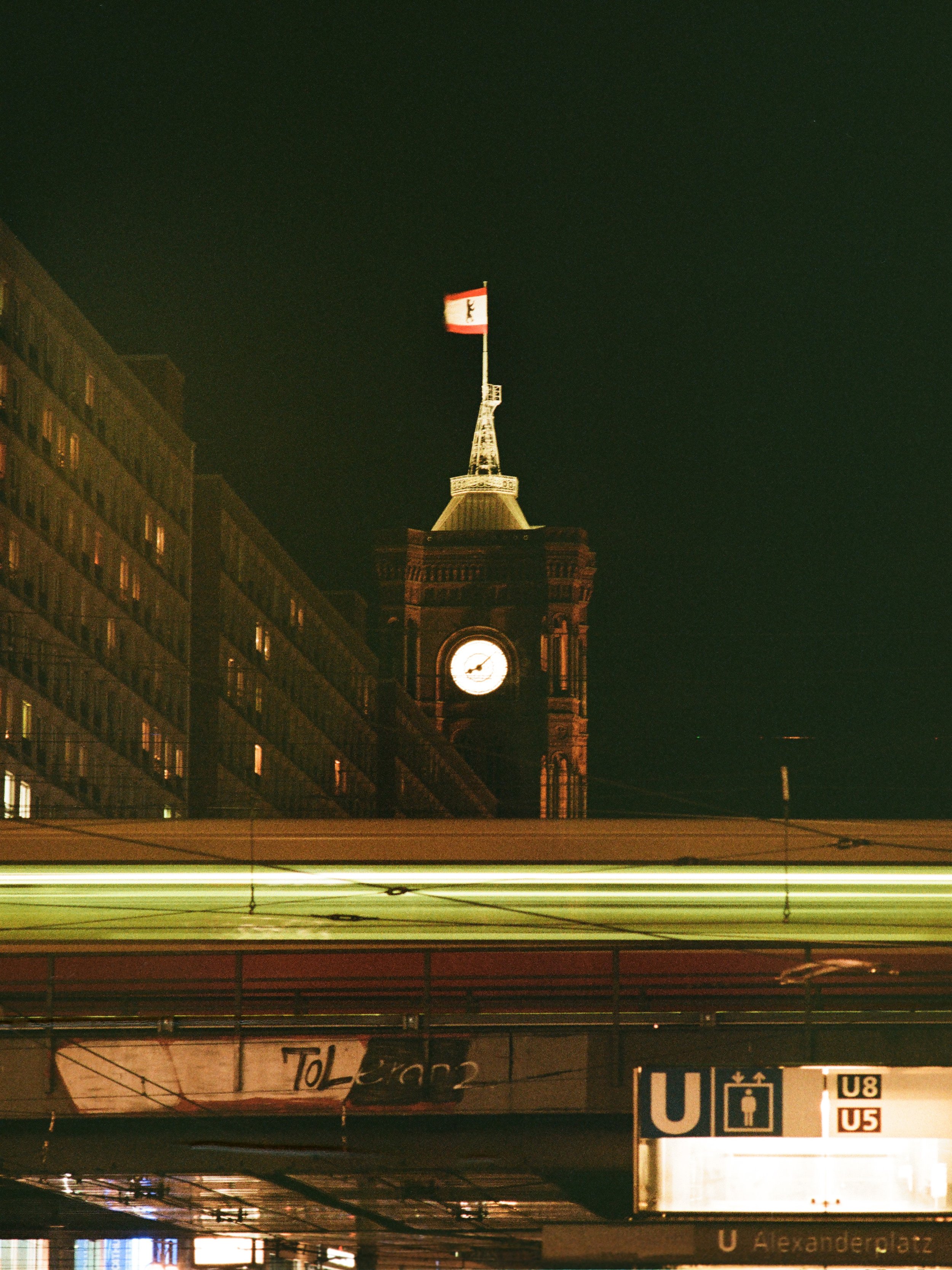 Ein dunkler Nachthimmel mit einem befeuerten Uhrturm, auf dem eine Deutschlandfahne weht, neben einem großen Gebäude in Berlin.