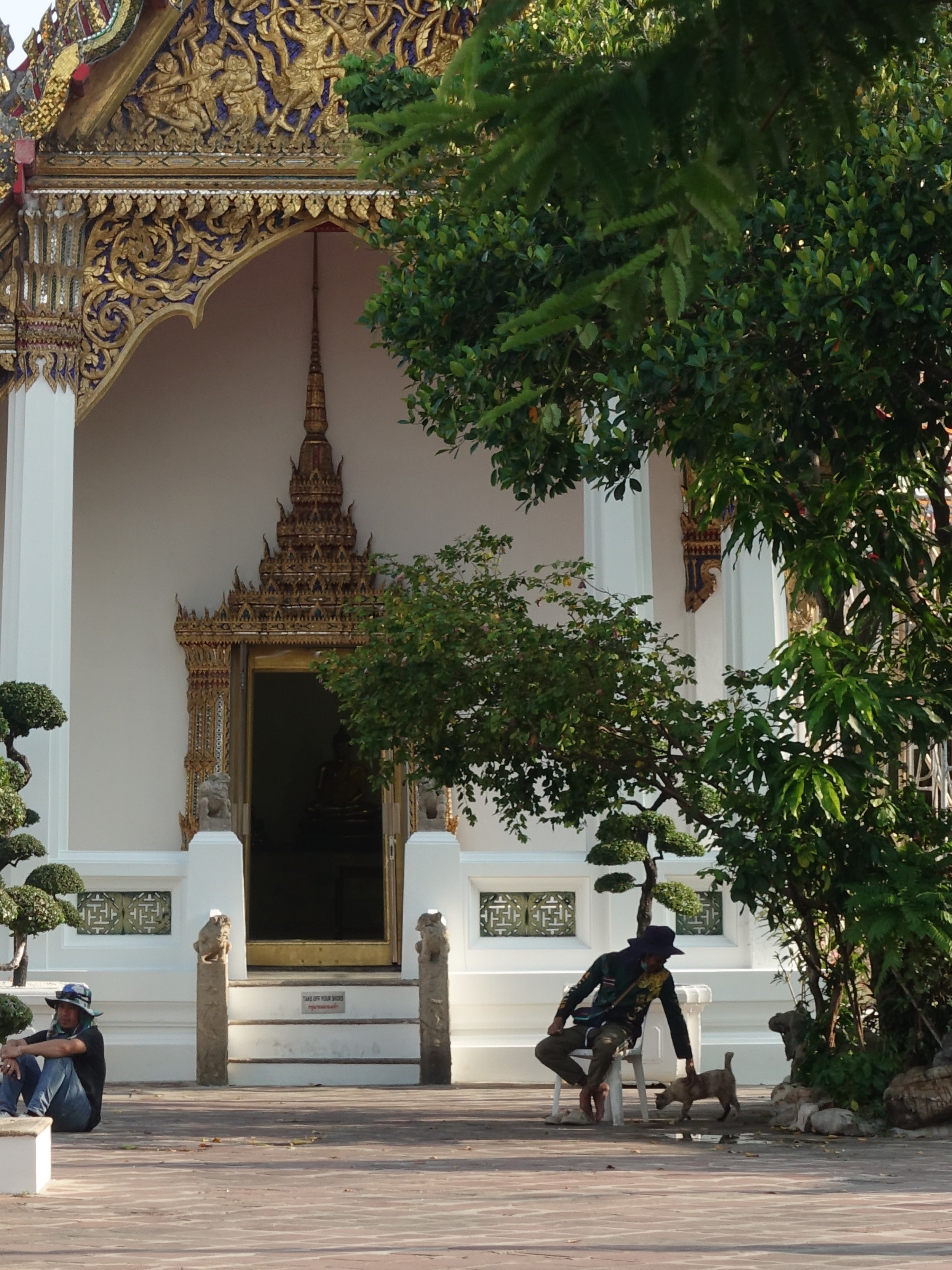 Ein Tempel, umgeben von Bäumen, mit einer goldverzierten Fassade und Stufen mit Steinstatuen. Zwei Männer sitzen im Schatten, einer streichelt eine Katze.