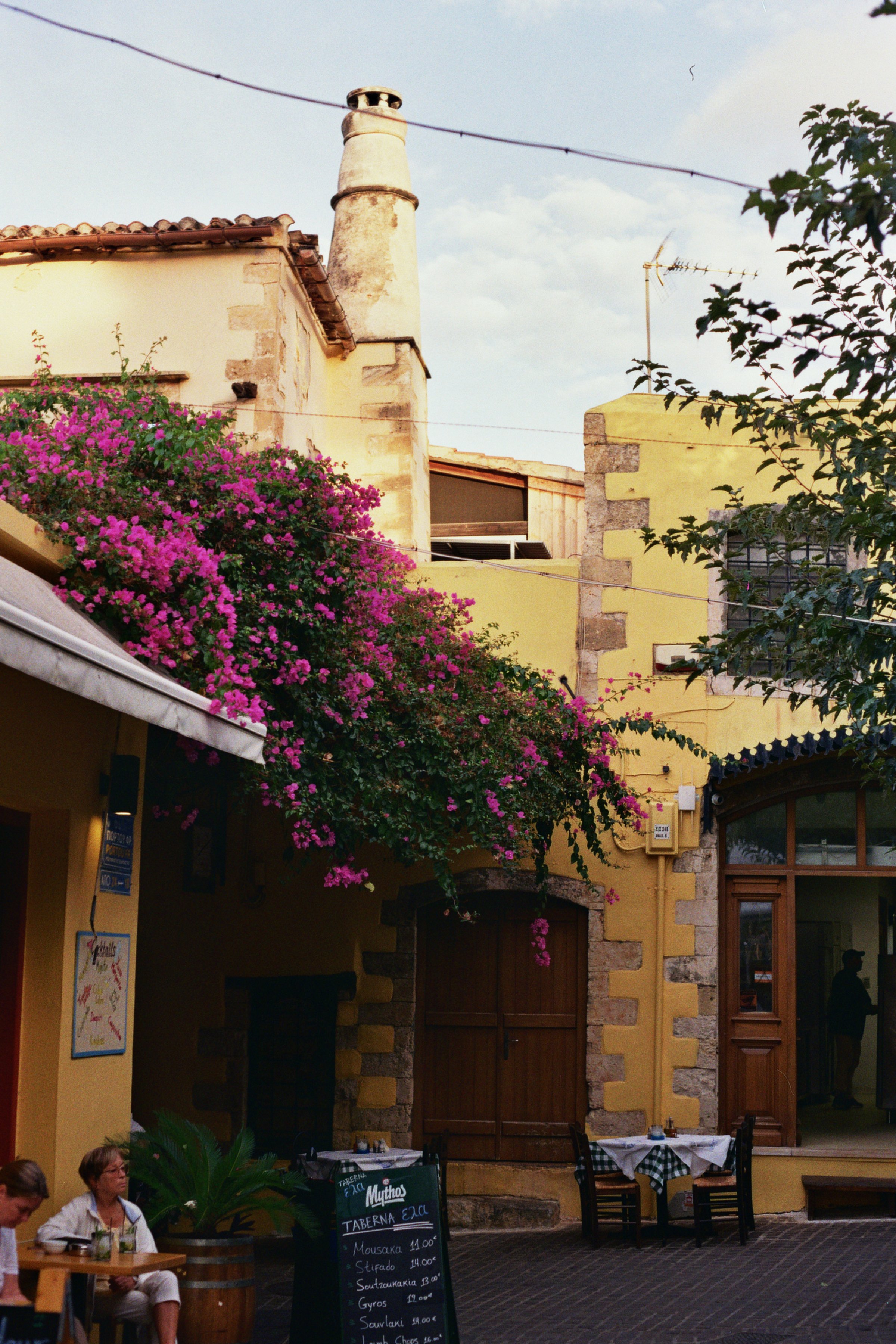 Straßenszene mit gelben Gebäuden, blühenden Bougainvillea, Tischen im Freien, und Menschen, die am Café sitzen, bei sonnigem Himmel.
