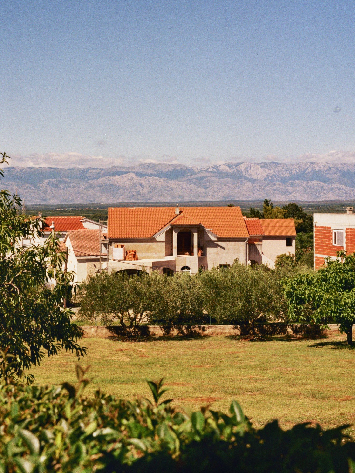 Ein Haus mit roten Ziegeldächern in einer grünen Landschaft, im Hintergrund Berge unter blauem Himmel.