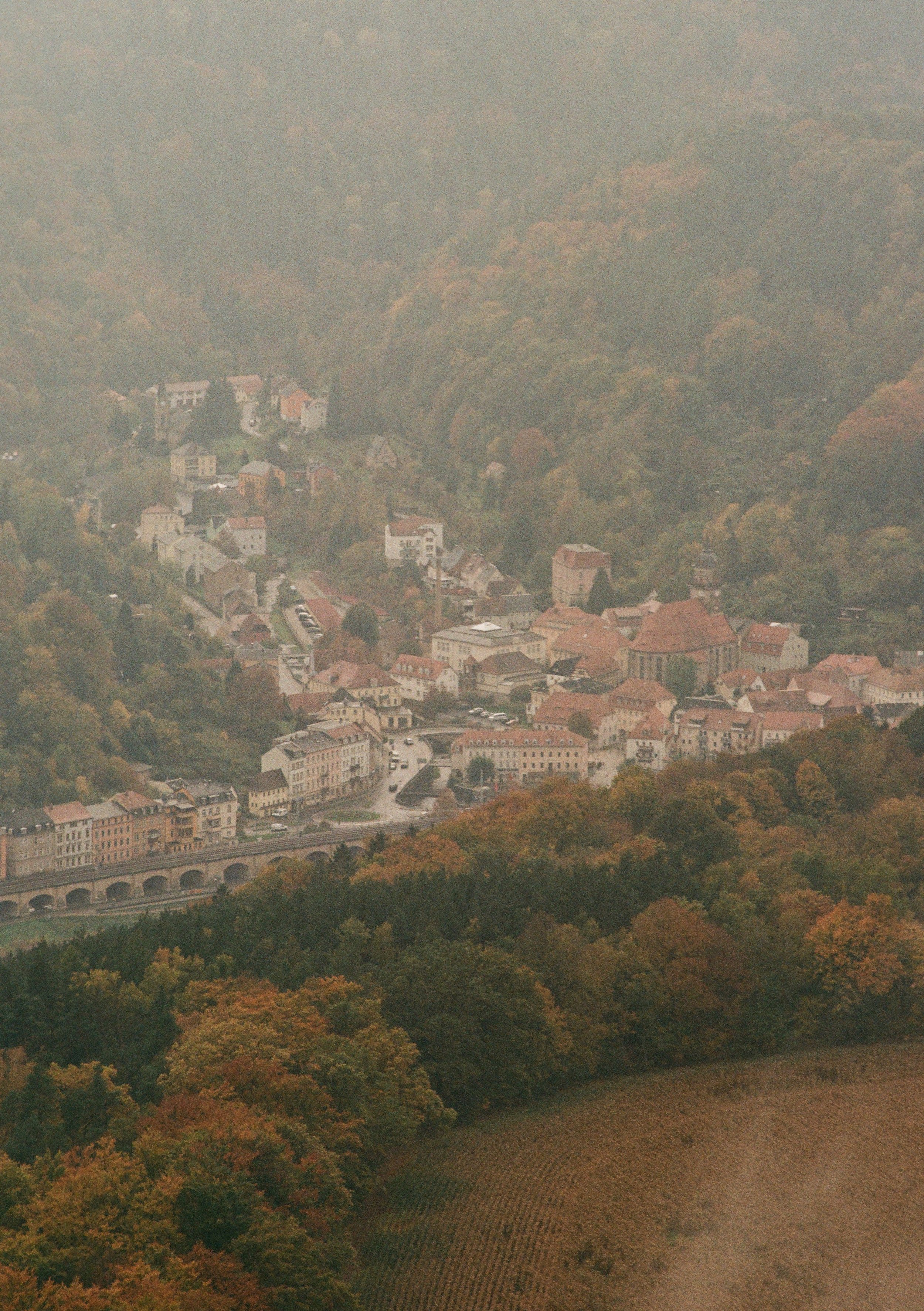 Ein malerisches Dorf inmitten von bewaldeten Hügeln im Herbst, mit roten und orangenen Laubbäumen, einige alte Gebäude und einen alten Fachwerkbrücken.