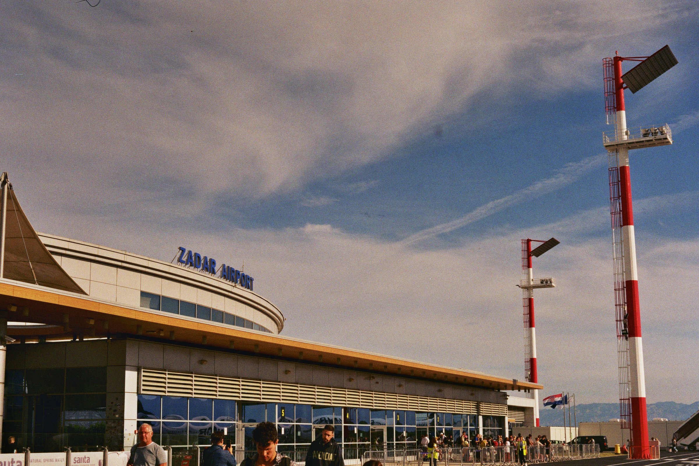 Eingang des Zadar Flughafens mit Menschen, und Zadar Airport Beschilderung unter einem bewölkten Himmel.