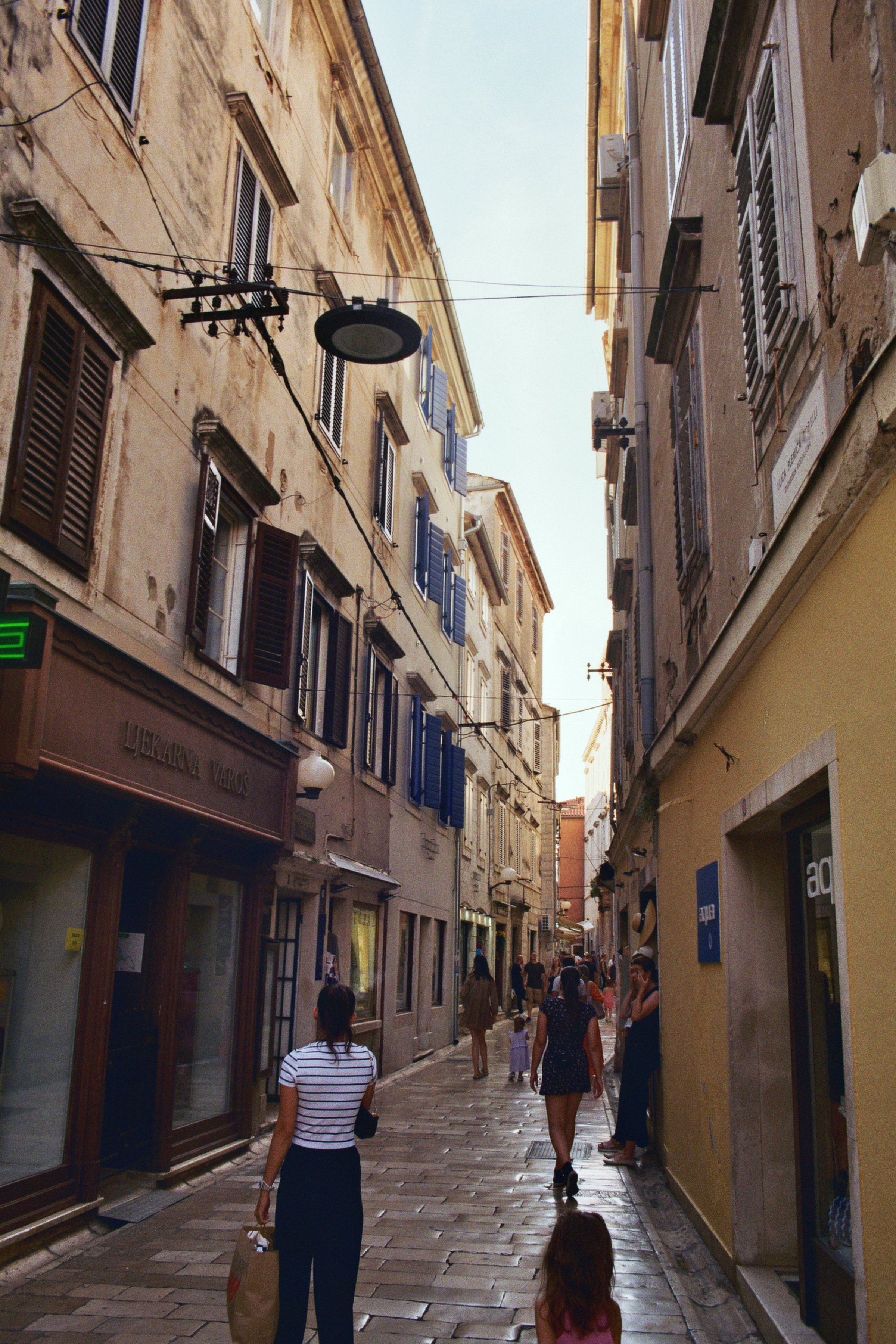 Blick auf eine enge, historische Stadtstraße mit verputzten Gebäuden, offenen Fensterläden und Fußgängern, die spazieren gehen.