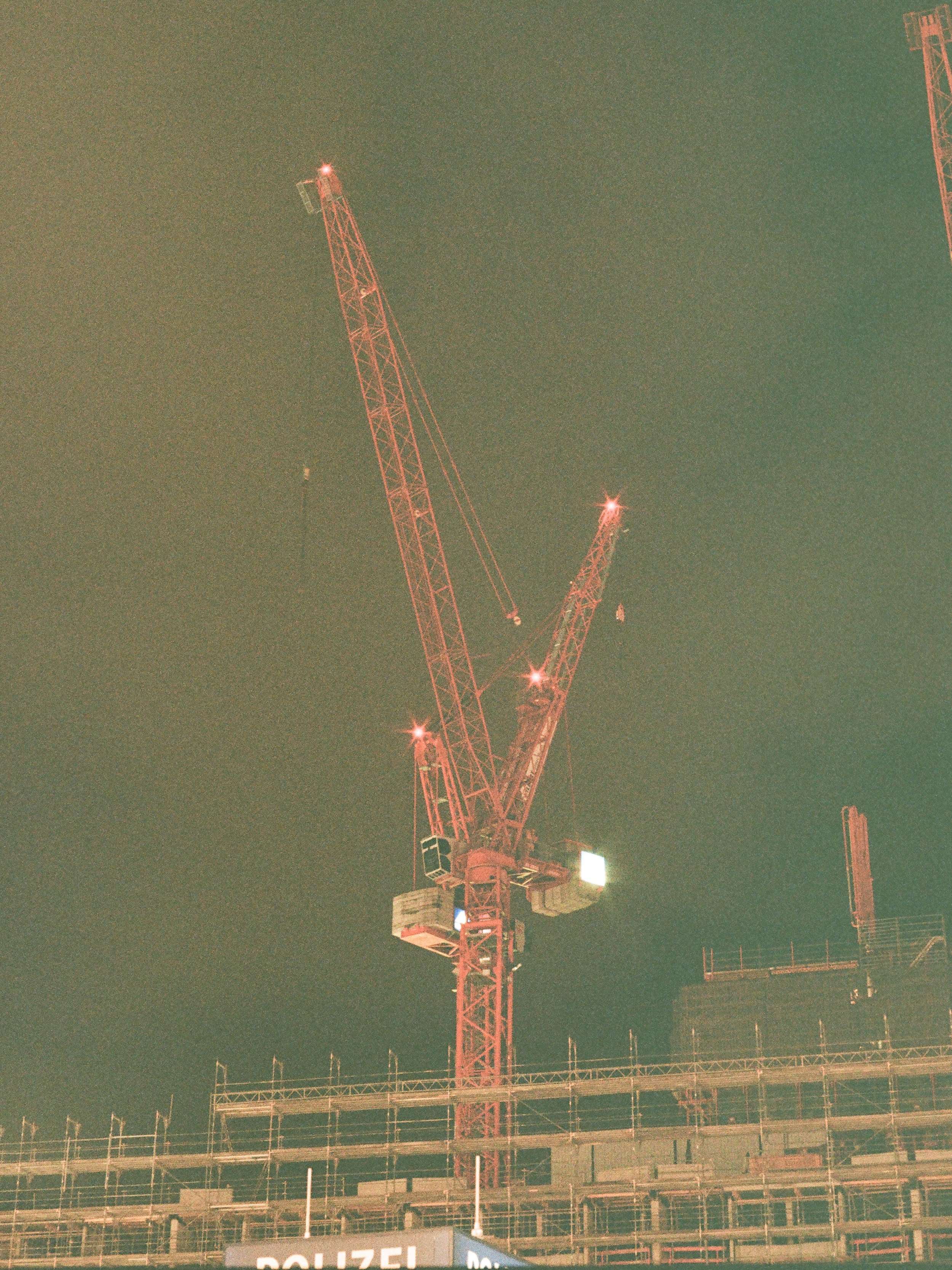 Baustellenkran bei Nacht, leuchtende Lichter, im Hintergrund eine Baustelle und ein Polizeibanner.