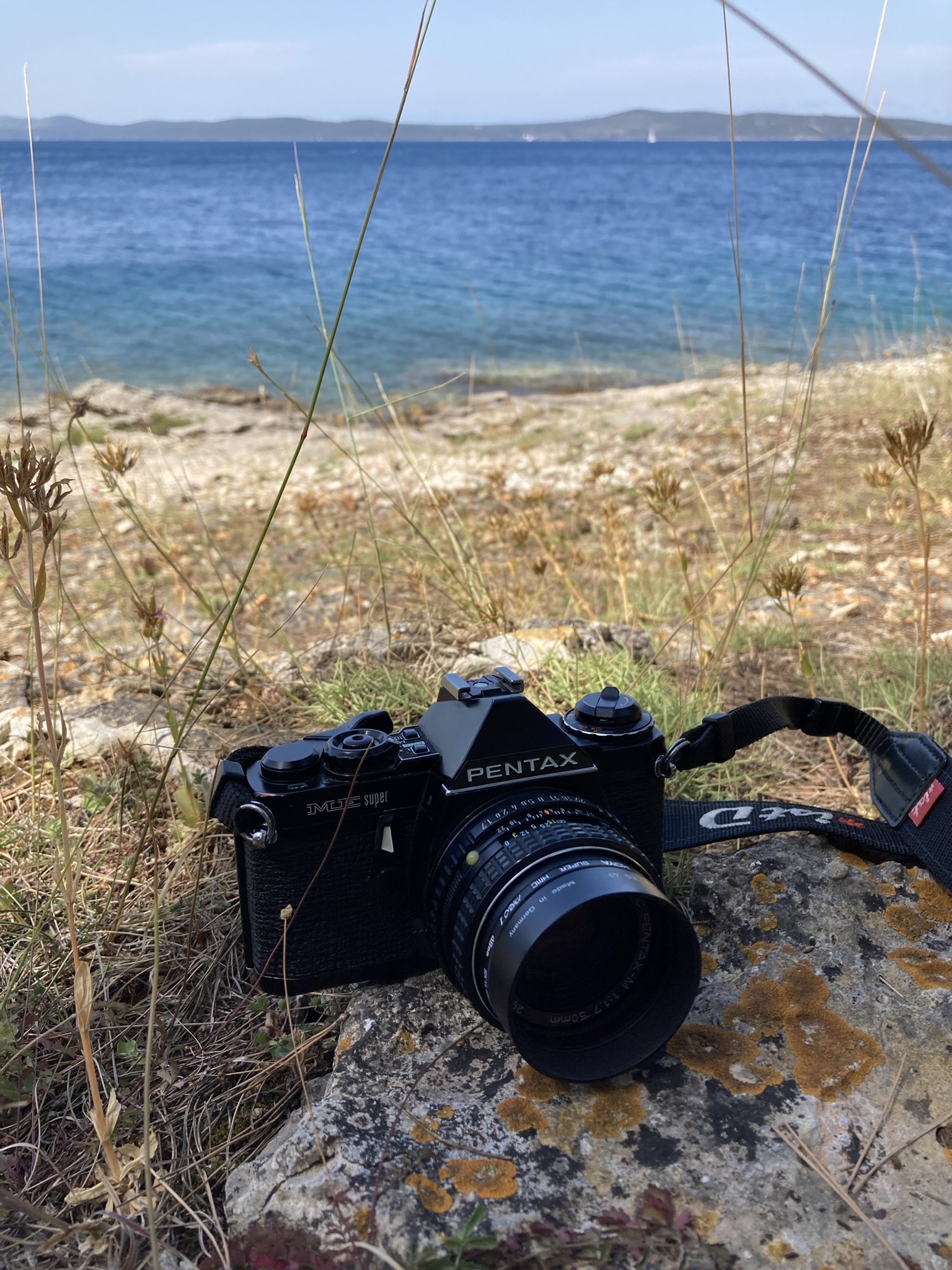 Eine Kamera liegt auf einem Stein am Strand, im Hintergrund das Meer und eine Insel bei klarem Wetter.