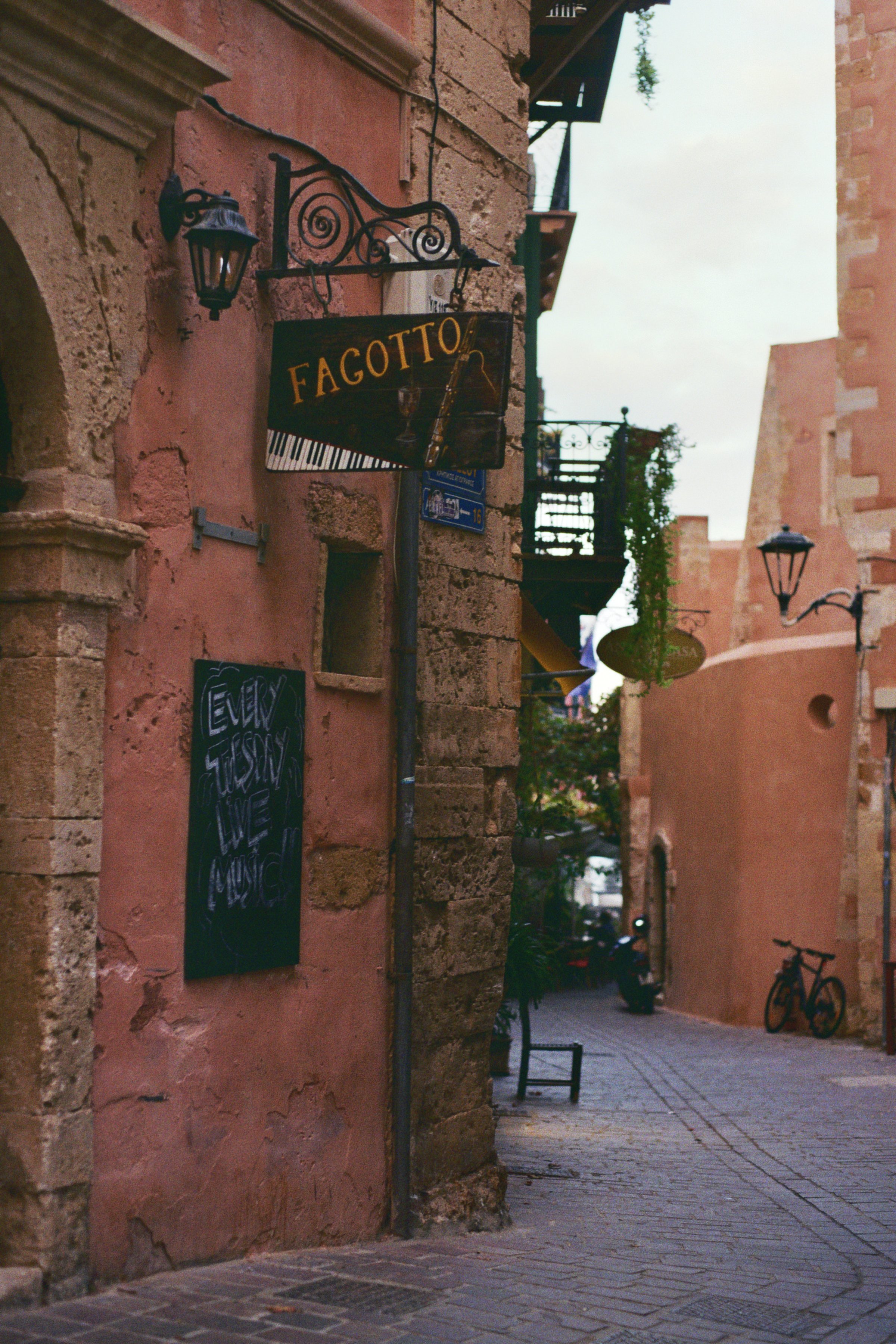 Ein enge Gasse mit alten, rotbraunen Gebäuden. Ein Schild mit der Aufschrift "FAGOTTO!" hängt an der Wand sowie eine Tafel mit der Aufschrift "Every Tijuana We Music". Es stehen einige Fahrräder an der Seite, und die Straßenlaternen sind an den Gebäu