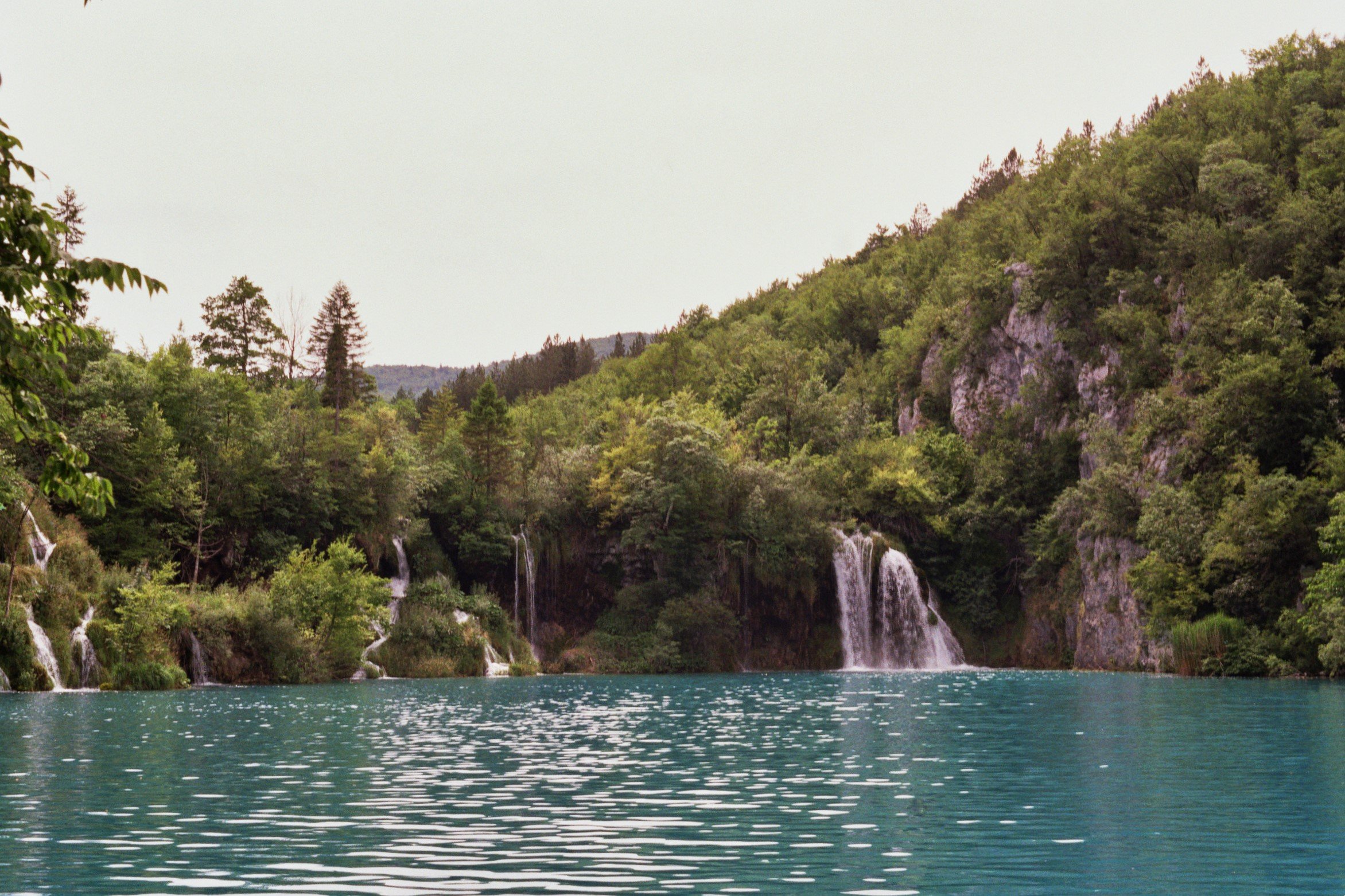 Blick auf einen klaren, blauen See mit kleinen Wasserfällen, umgeben von einem grünen, bewaldeten Hügel