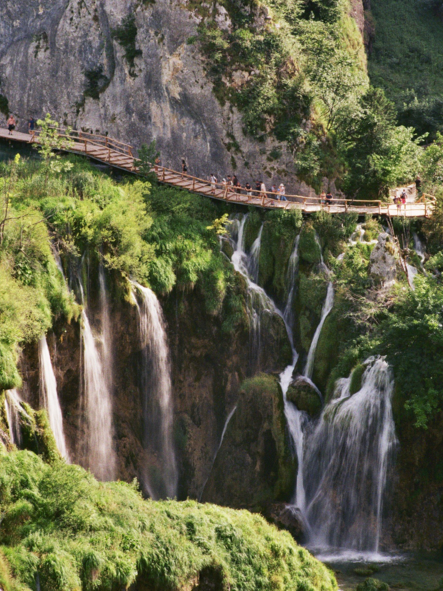 Wanderweg entlang eines Wasserfalls in einer grünen Berglandschaft, mit mehreren Personen, die den Weg entlang gehen.