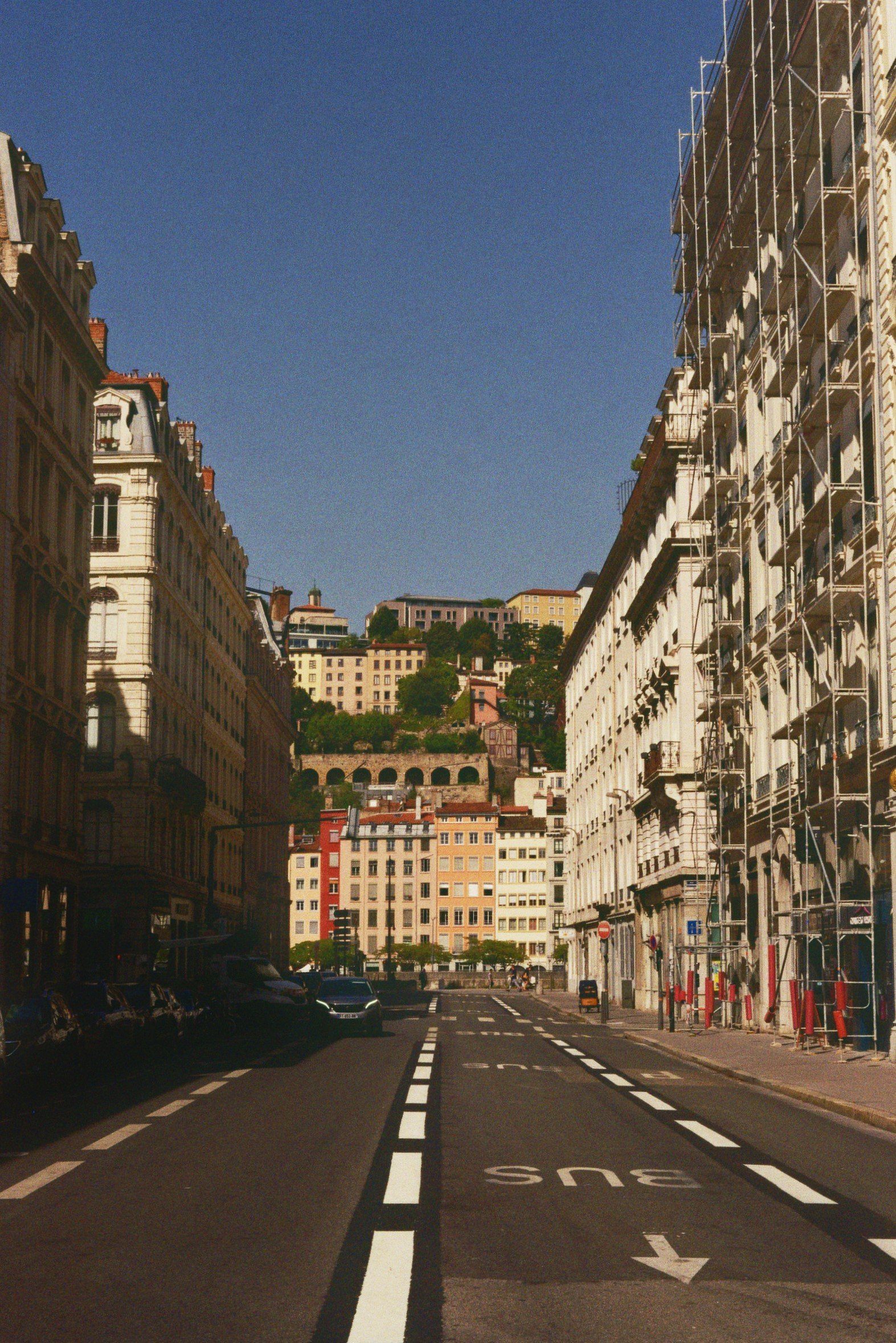 Stadtstraße mit umliegenden Gebäuden, Baustellengeräten und einem gelben Bus, im Hintergrund Hügel mit weiteren Gebäuden, blauer Himmel.